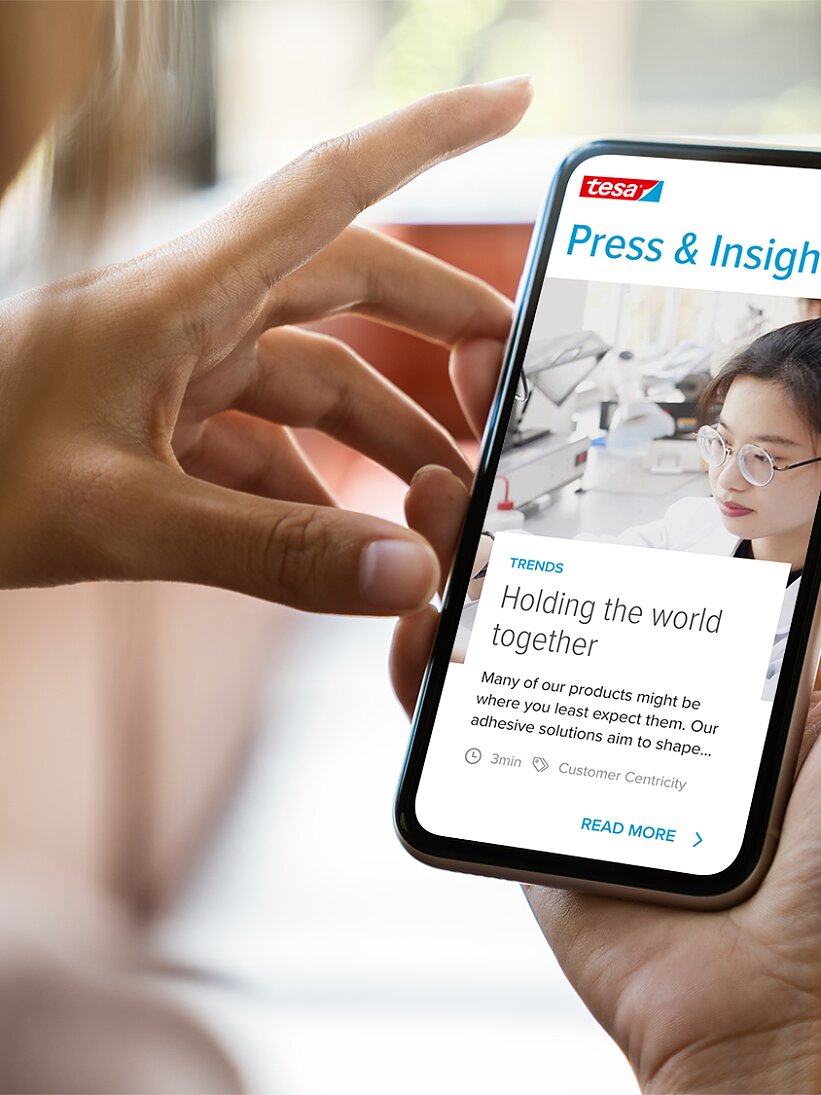 A person holding a smartphone displaying a webpage titled Press & Insights. The screen shows an article with an image of a woman behind a microscope and text about tesa tape adhesives. The smartphone is held in a hand with a blurred background. (This text has been generated by AI)