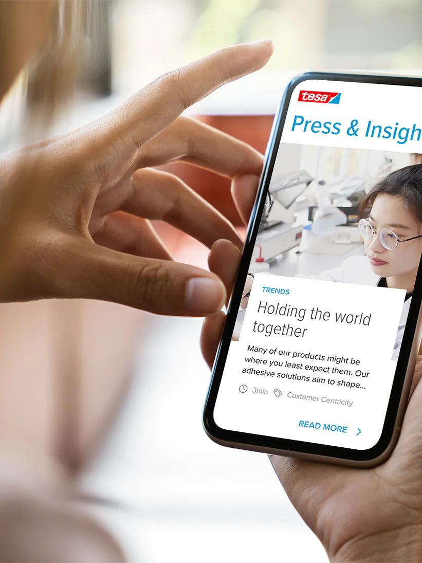 A person holding a smartphone displaying a webpage titled Press & Insights. The screen shows an article with an image of a woman behind a microscope and text about tesa tape adhesives. The smartphone is held in a hand with a blurred background. (This text has been generated by AI)