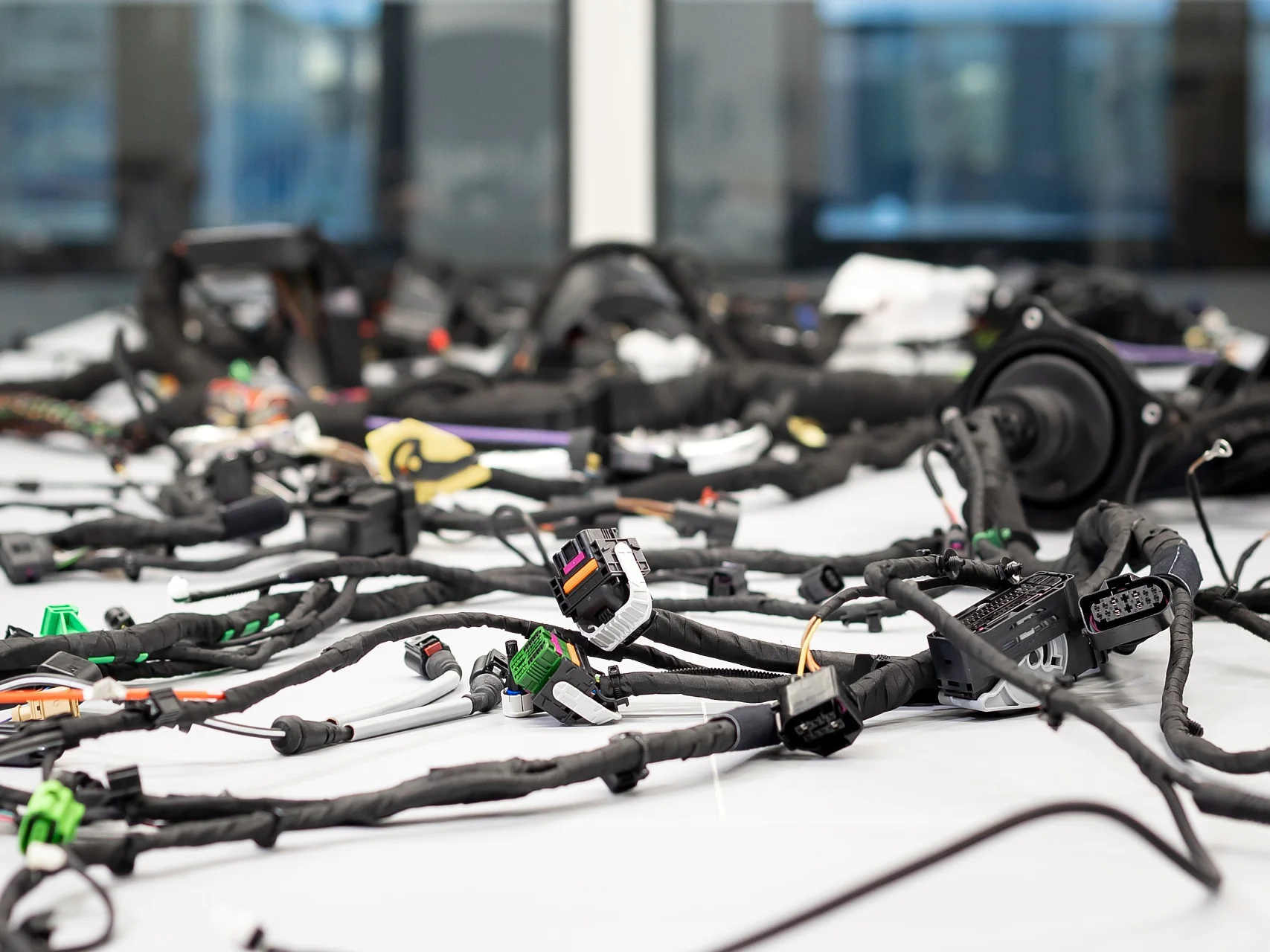A large number of electronic cables and connectors are spread across a white table. The background shows blurred glass windows and a building exterior. The setup appears to be in a work or testing area for electronics, where tesa tape is likely used to secure or organize the cables effectively. (This text has been generated by AI)