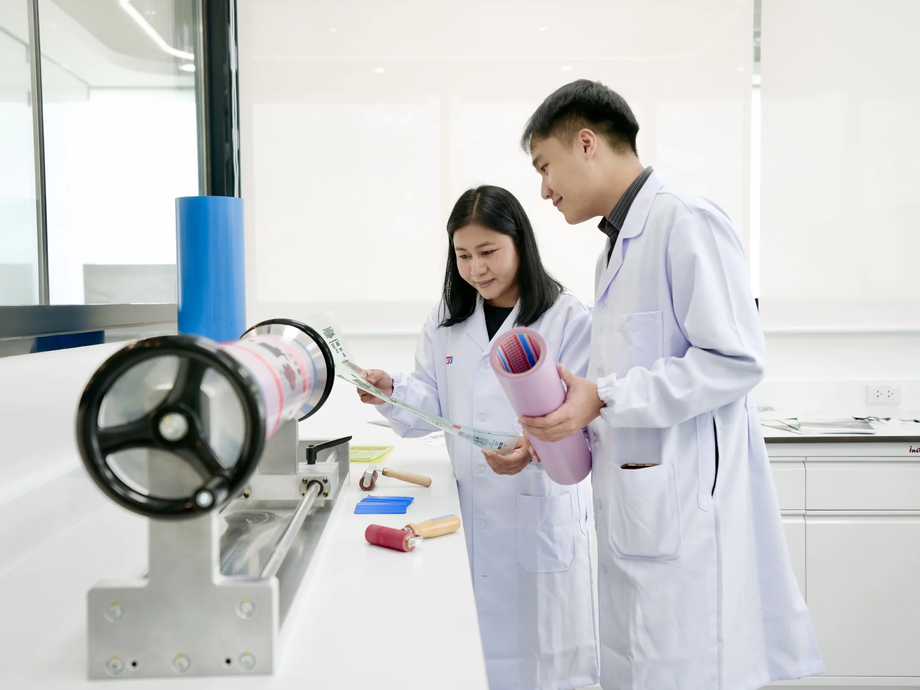 Dua orang dengan jas lab memeriksa lembaran bahan dan dokumen, termasuk pita tesa, di samping mesin di laboratorium yang terang. (This text has been generated by AI)