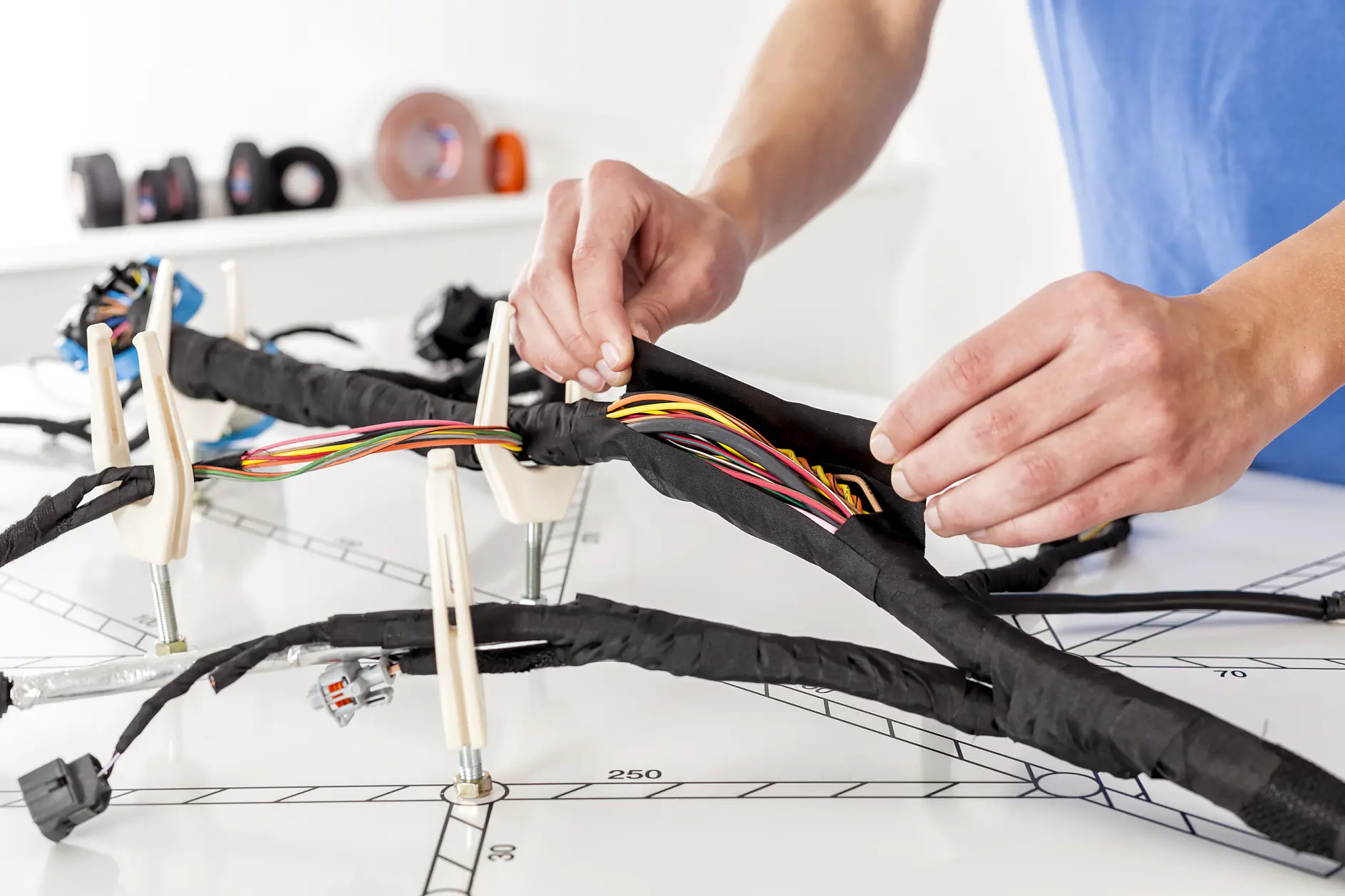 Hands wrapping black electrical tape around a bundle of colorful wires on a white workbench indoors. (This text has been generated by AI)