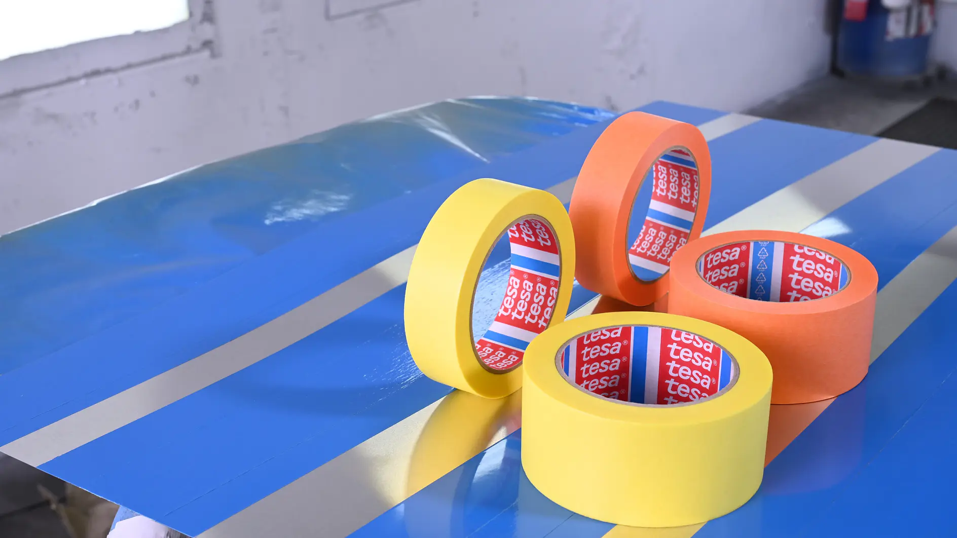 Four rolls of tesa adhesive tape in yellow and orange on a blue surface with reflective silver stripes indoors.