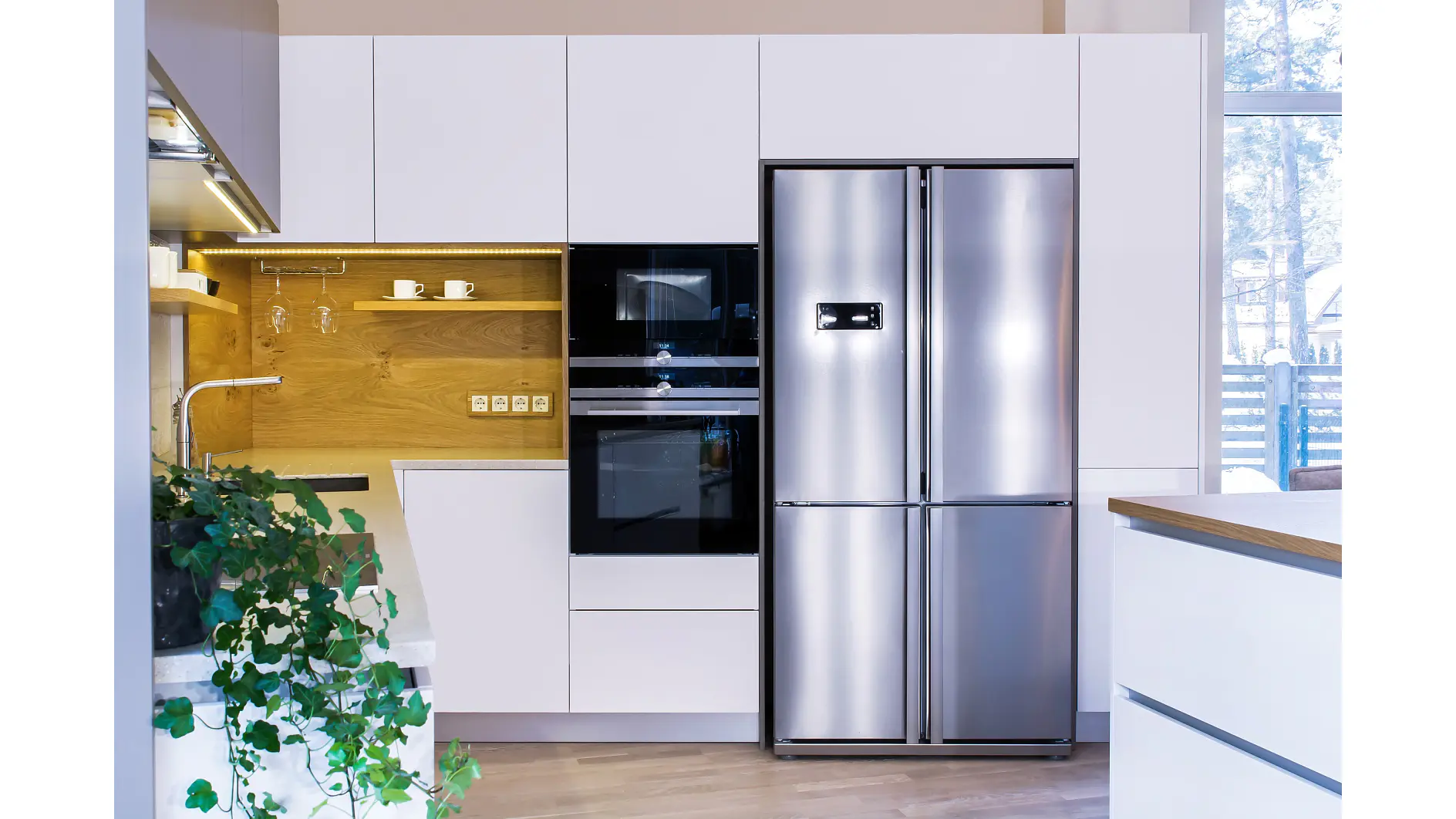 Modern kitchen with white cabinets, built-in black oven and microwave, wooden backsplash with open shelves secured using tesa tape, and a large stainless steel double-door refrigerator. A green plant is visible on the countertop. (This text has been generated by AI)