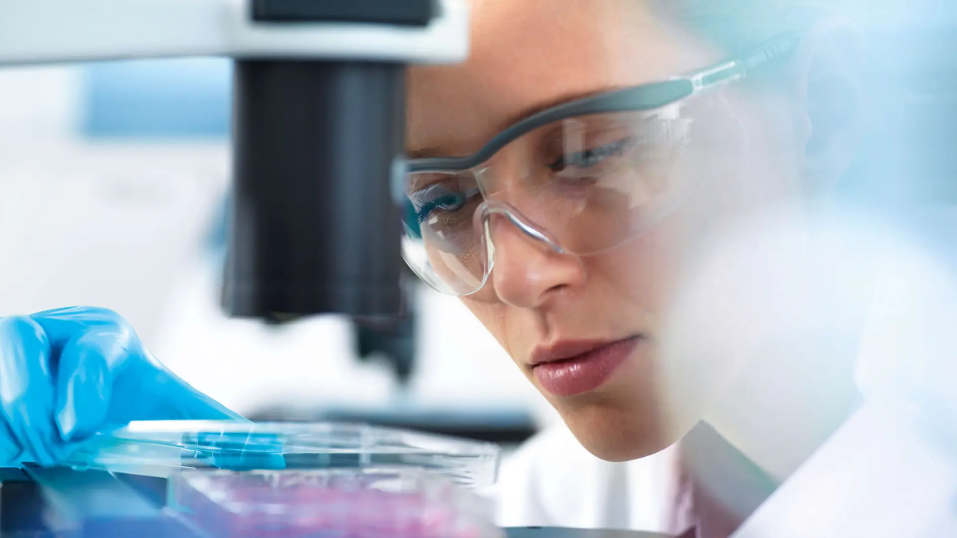 A person in safety goggles and gloves closely examines a sample under a microscope. The background is blurred, emphasizing the scientific laboratory setting.