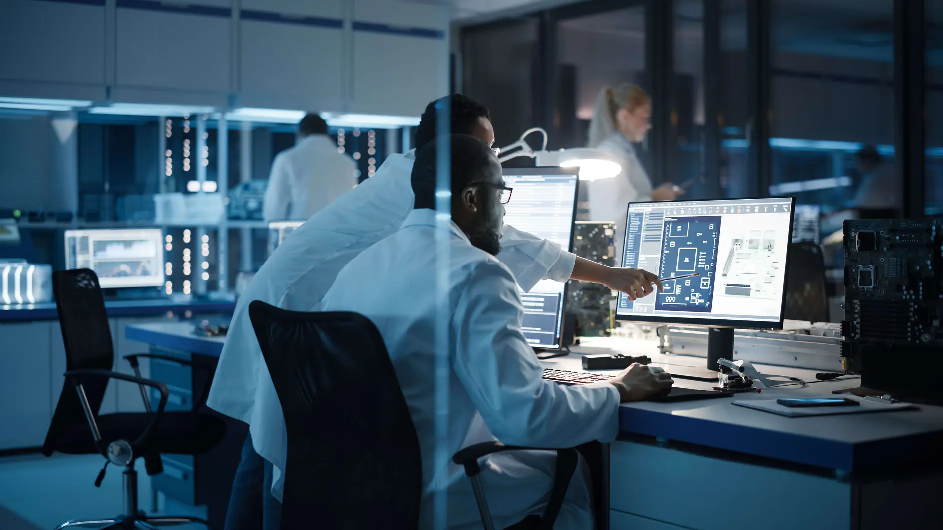 Two scientists in lab coats work at a computer that displays complex circuit diagrams on the screen. One points at the monitor while the other sits, observing. The lab is equipped with electronic equipment, and there are more researchers working in the background.