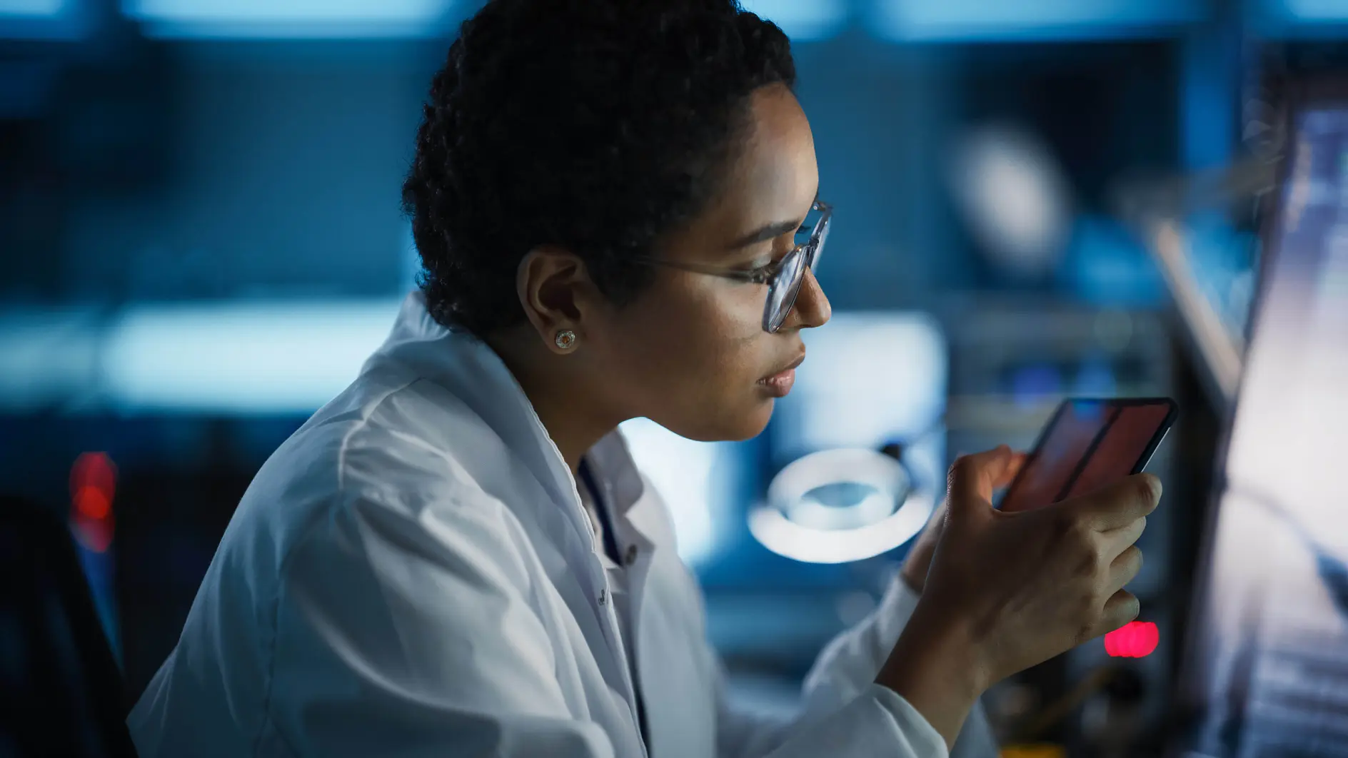 A person wearing a white lab coat and glasses is sitting in a lab environment, closely examining a smartphone secured with tesa tape. The background is blurred, featuring computer screens and lab equipment. The lighting is dim, highlighting the focus on the smartphone wrapped with tesa tape.
