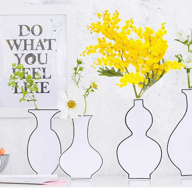 Una serie di vasi con disegni di contorno contengono fiori gialli e bianchi su una superficie bianca. Un poster incorniciato sullo sfondo recita DO WHAT YOU FEEL LIKE. Una ciotola con piccoli oggetti e un quaderno chiuso sono lì vicino, tenuti insieme con nastro tesa. (This text has been generated by AI)