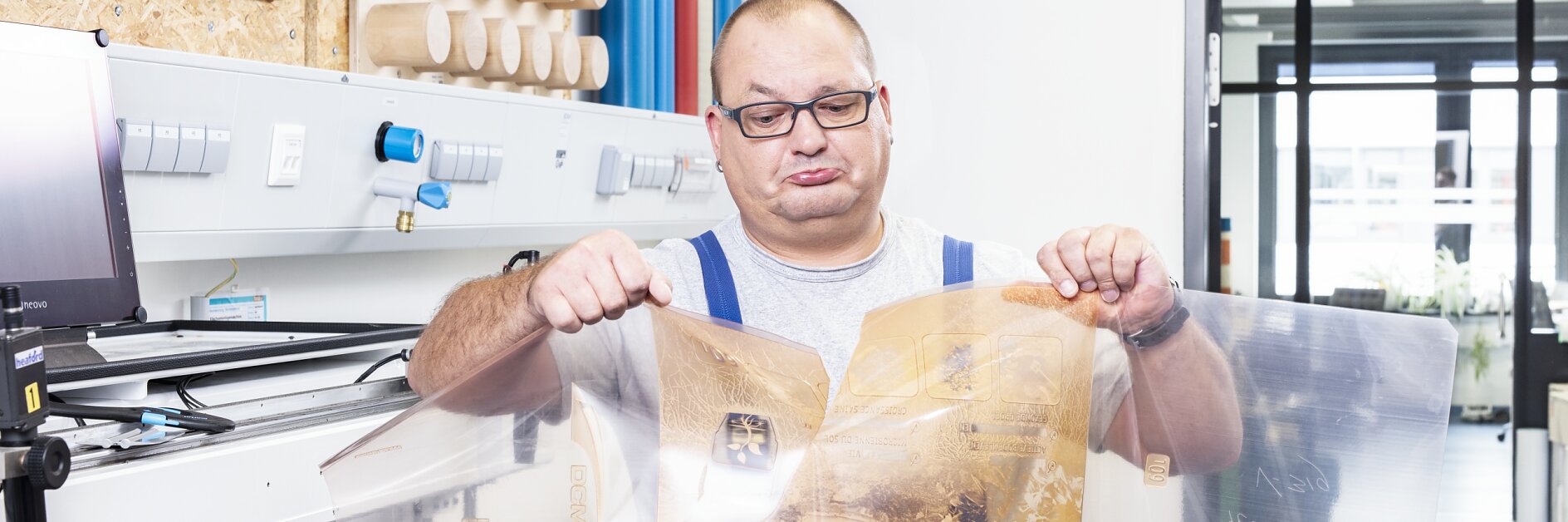 A man wearing glasses and blue overalls inspects transparent plastic sheets with printed designs in an industrial setting. Behind him are wooden rollers and machinery used for processing tesa tape. A window reveals a modern office space in the background.