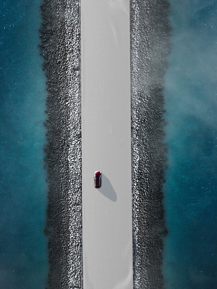 Aerial view of a red car driving on a narrow, straight road flanked by blue water on both sides. The road is elevated, with rocky edges lining the sides. The water appears calm with a slight mist or fog hovering above it. (This text has been generated by AI)
