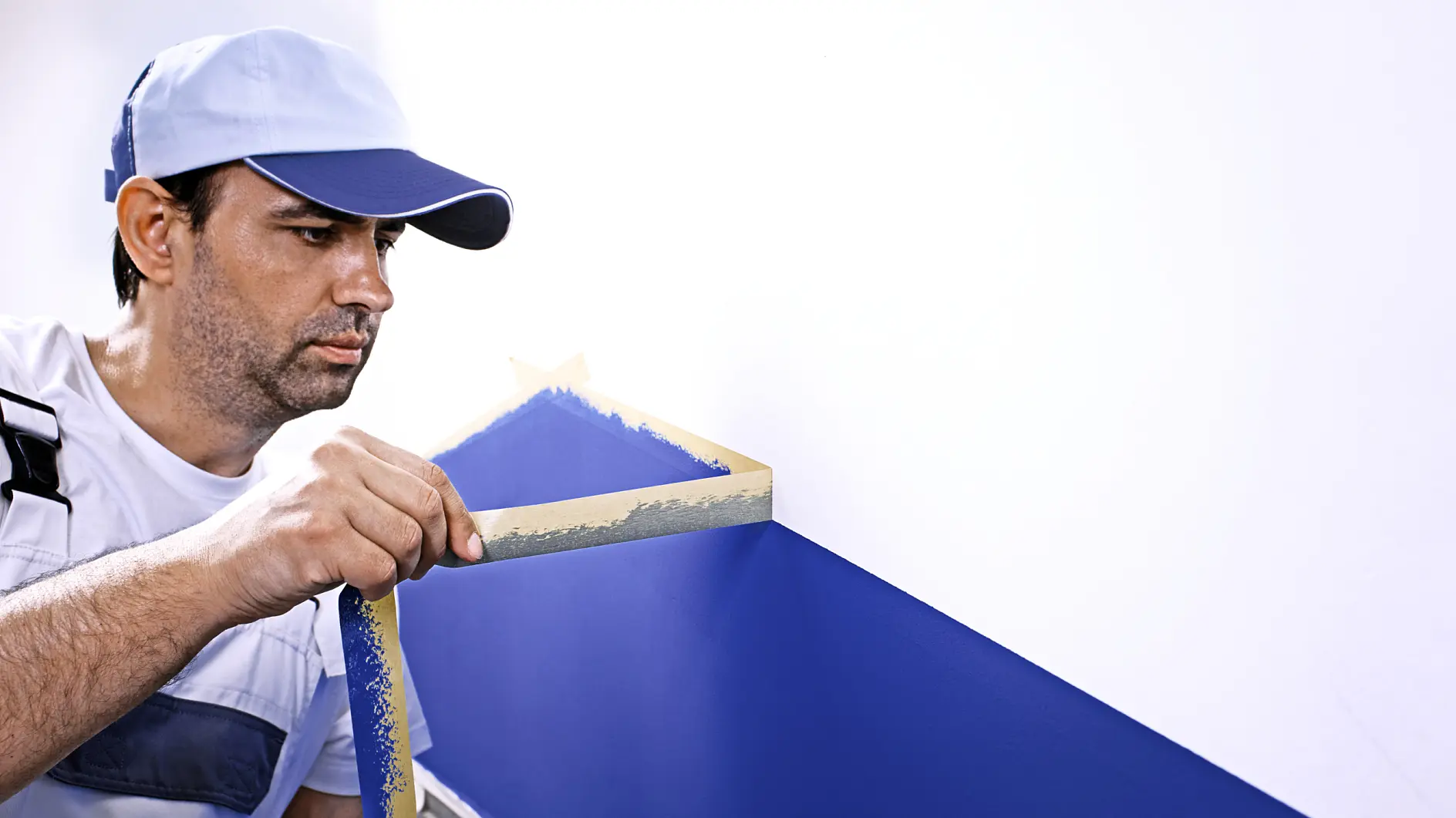 A man wearing a blue cap and a white shirt with blue overalls is painting a geometric design on a wall. He is peeling tesa tape from a blue-painted section on a white wall. A paint can is visible at his side.