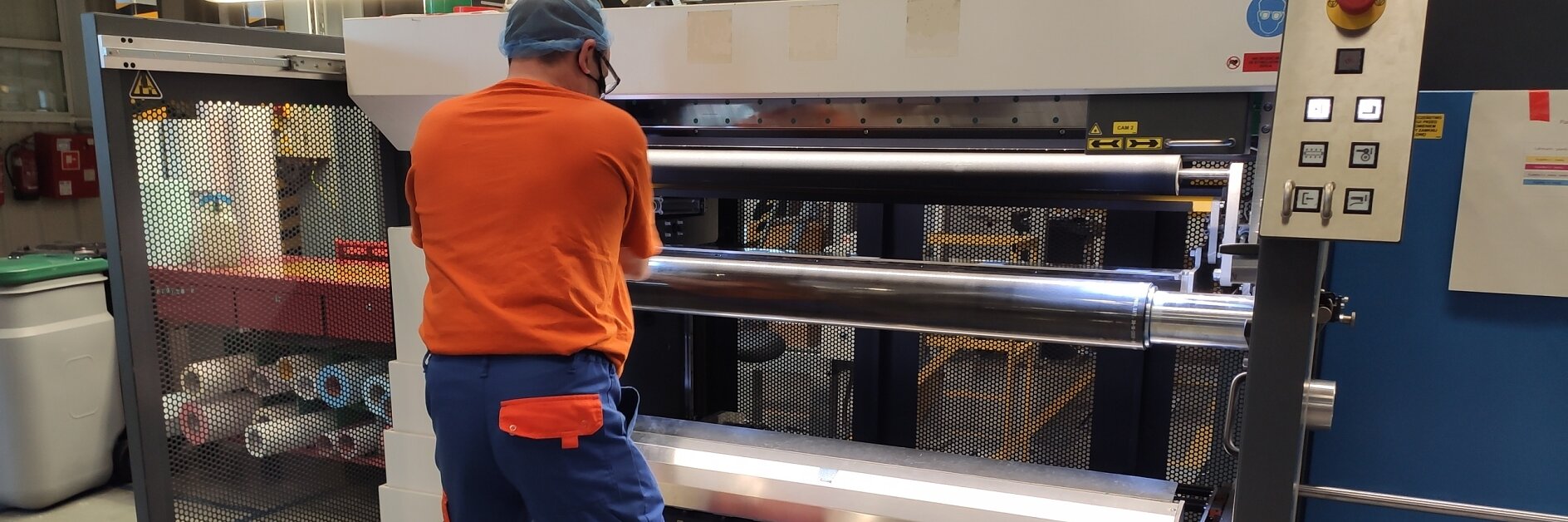 A worker in an orange shirt and blue pants operates a large industrial machine with rollers in a factory setting. He is facing the machine, which has a control panel at the top. Various materials, including tesa tape, are visible in the background.