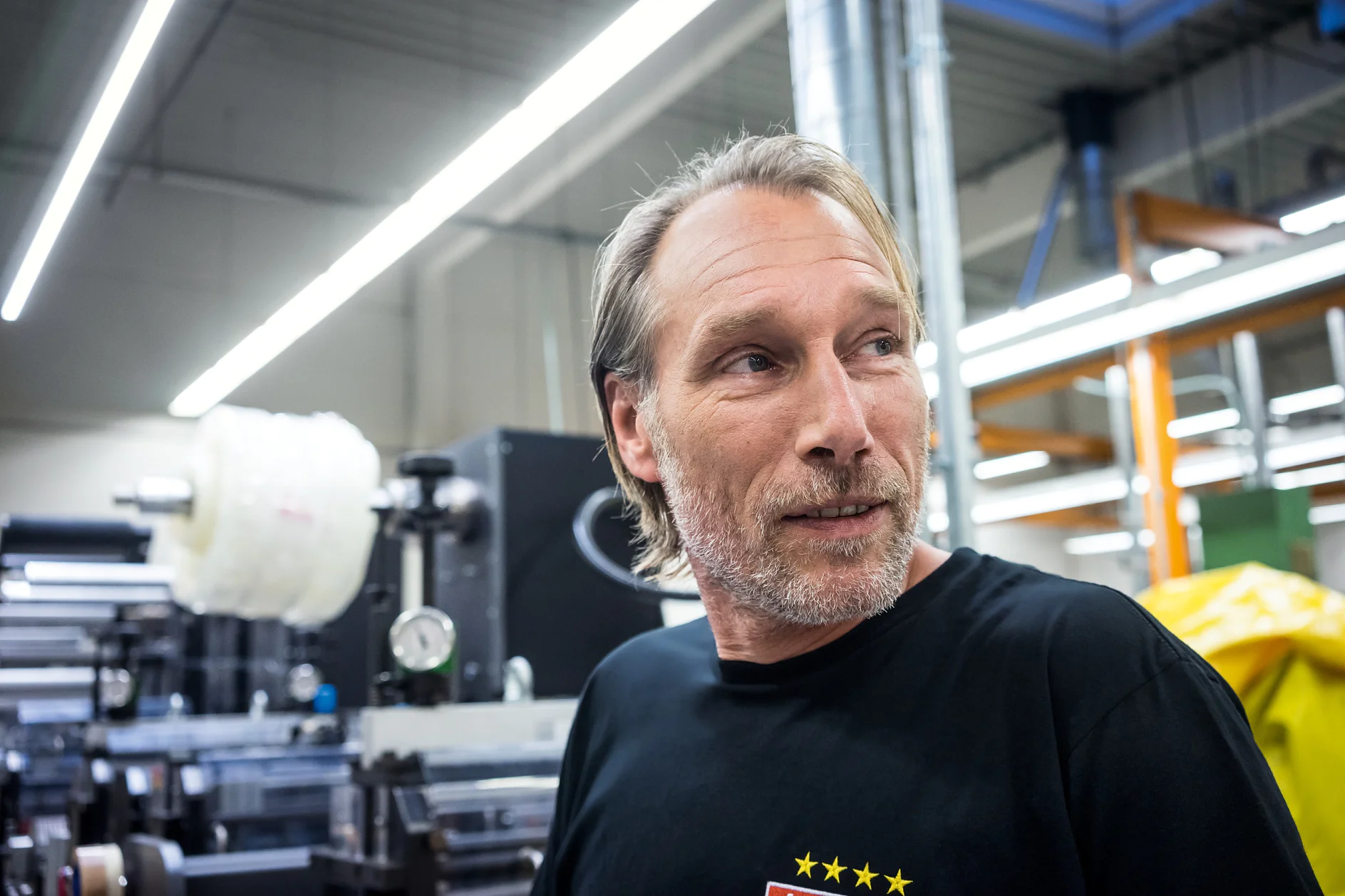 A man with light brown hair and facial hair stands in an industrial setting with machinery in the background. He is wearing a black T-shirt with yellow stars on it. Bright overhead lights illuminate the area, and several rolls of tesa tape can be seen nearby. (This text has been generated by AI)