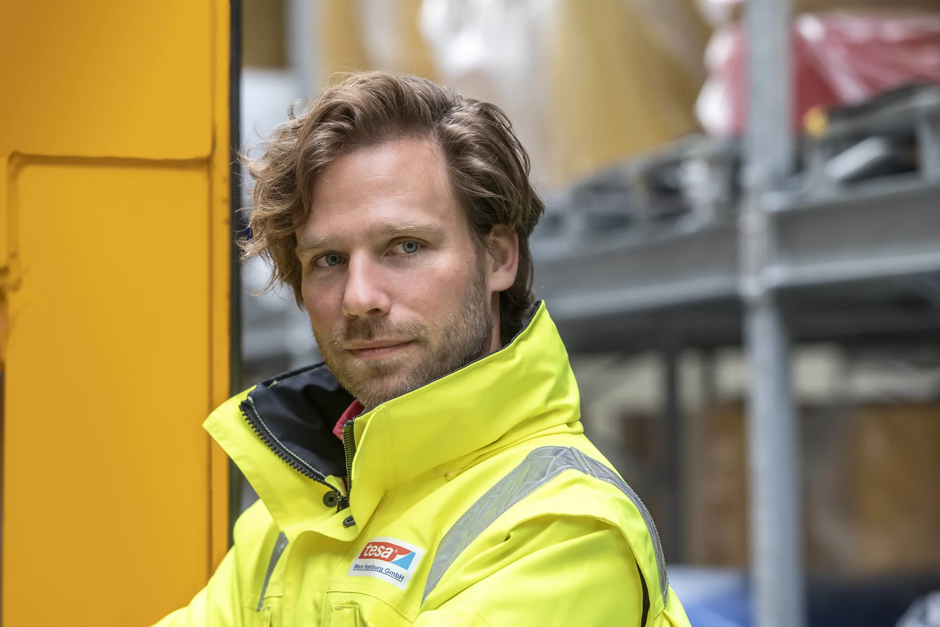 A person with wavy hair is wearing a bright yellow high-visibility jacket. They stand next to a yellow object in an industrial setting. Shelving with various covered items is visible in the background, all secured using tesa tape. (This text has been generated by AI)