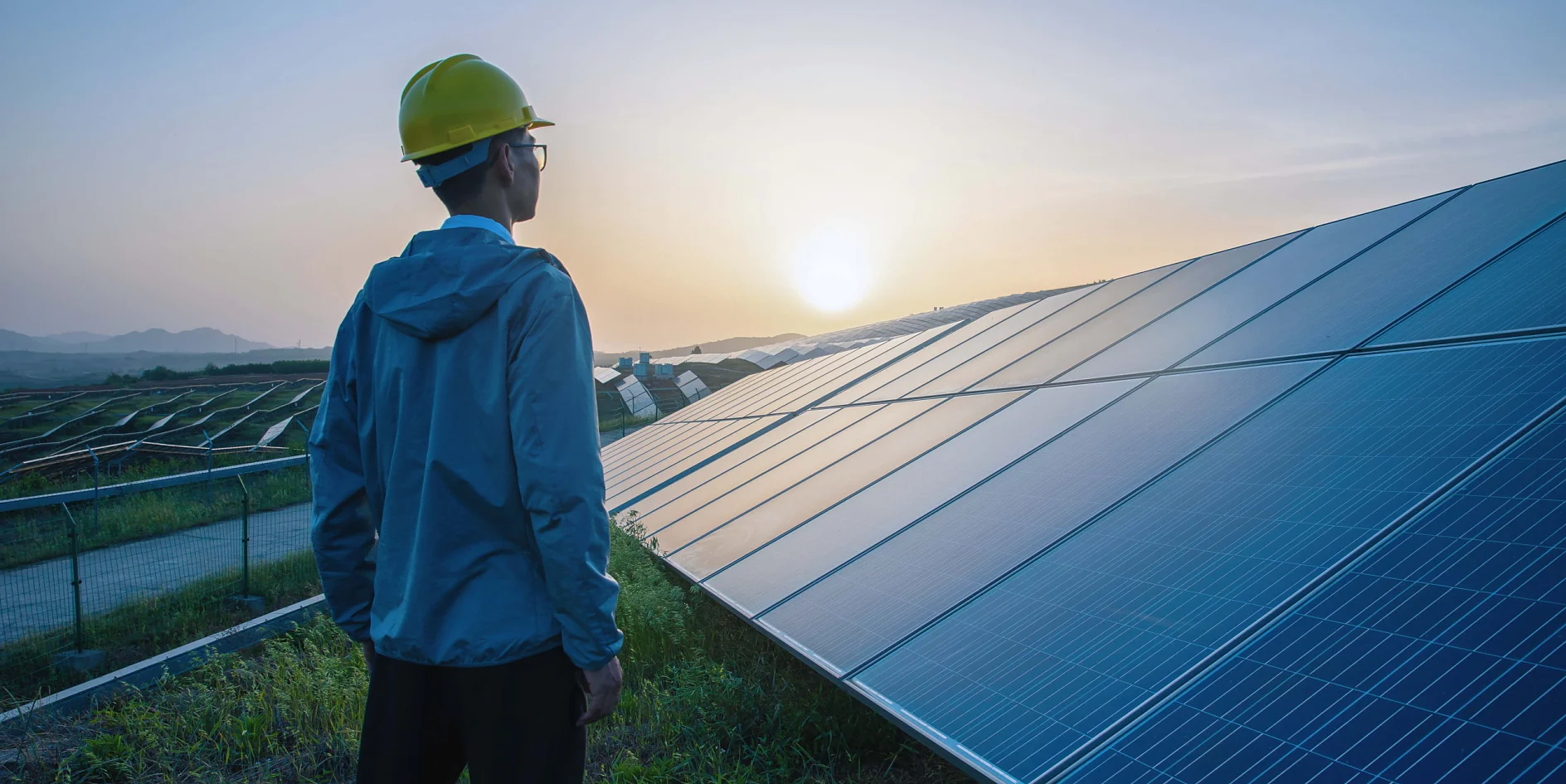A person in a yellow hard hat and blue jacket stands in a grassy area, looking at a large array of solar panels at sunset. The sky is a gradient of orange and blue, and distant hills are visible under the setting sun. (This text has been generated by AI)