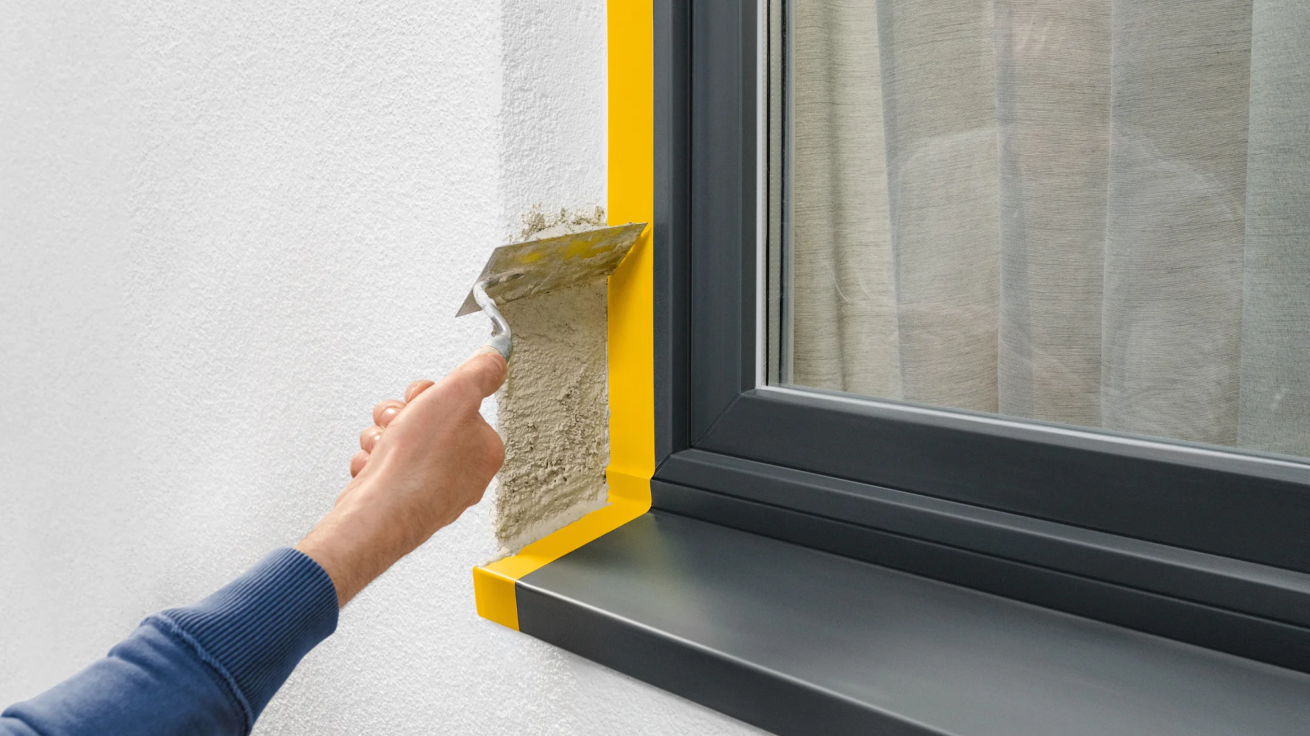Una persona usa una cazzuola per applicare lintonaco su un muro vicino a una finestra con una cornice scura. La finestra ha del nastro tesa giallo lungo i bordi e un riflesso della persona è visibile nel vetro. (This text has been generated by AI)