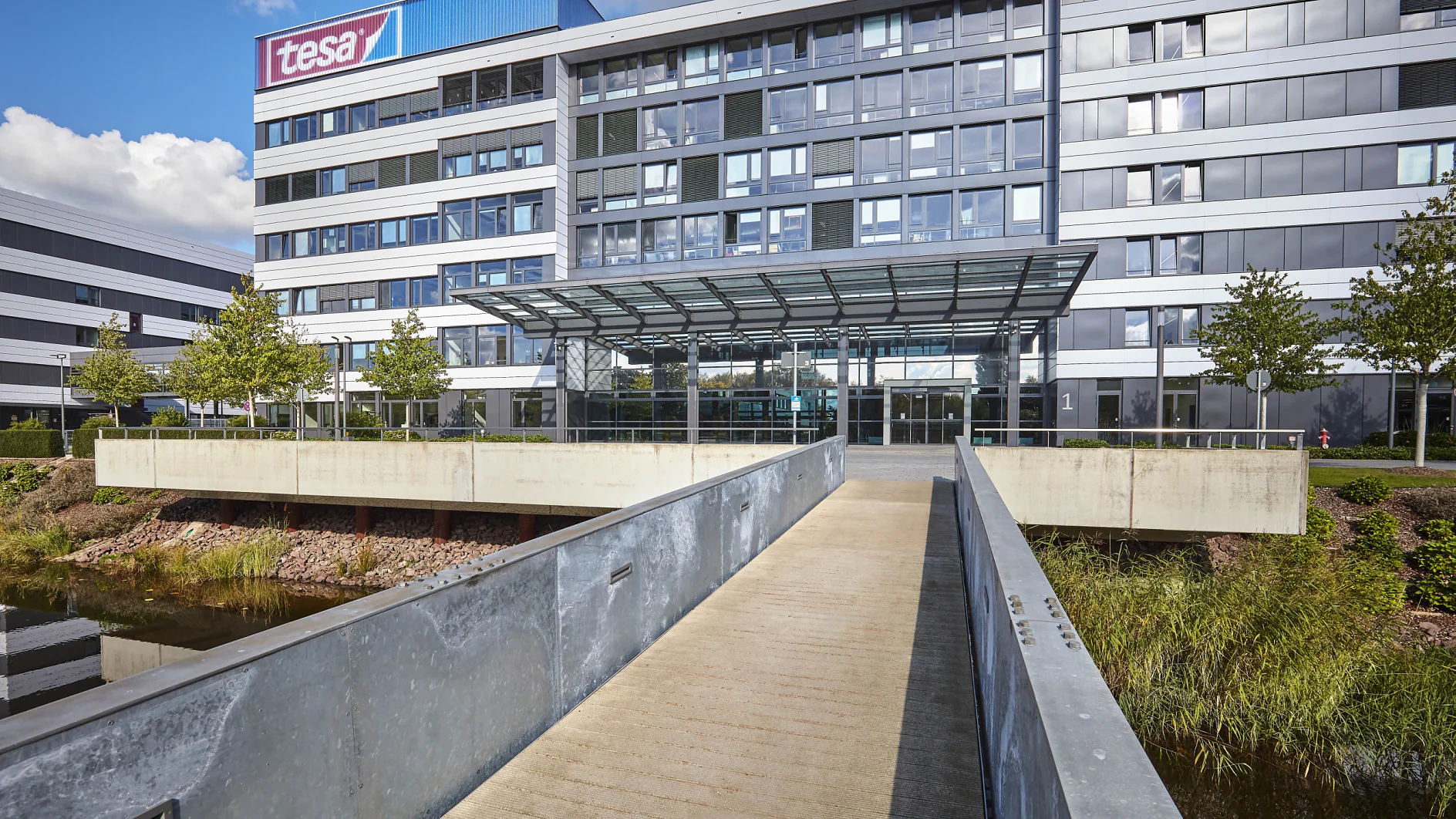 Exterior view of a modern office building with a glass and metal facade. A pedestrian bridge leads to the main entrance, featuring a glass canopy. The building is surrounded by greenery and water, and a tesa tape logo is visible at the top. (This text has been generated by AI)
