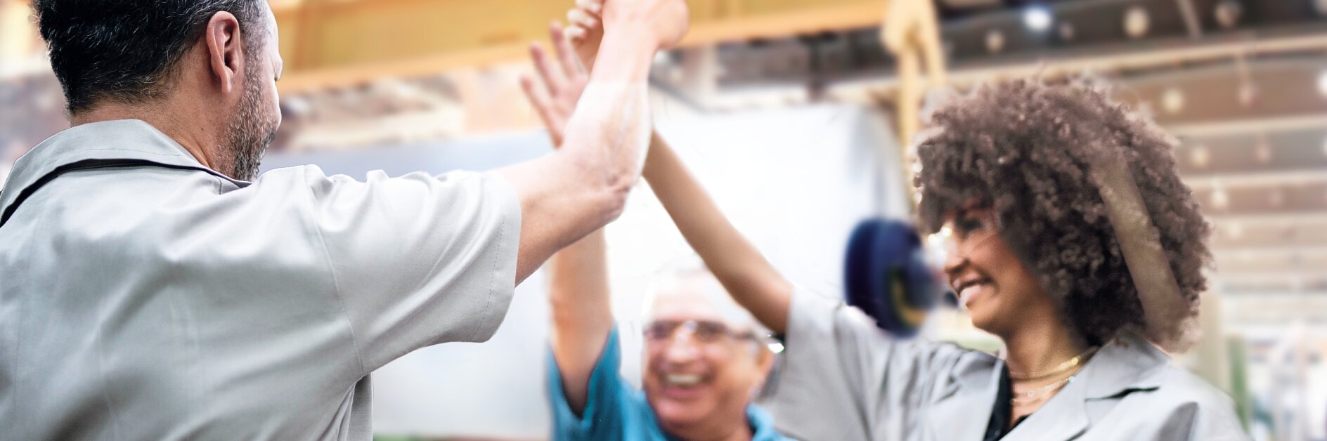 Three factory workers in uniforms celebrate inside an industrial setting, smiling and giving a high-five. The background shows machinery and bright lighting, along with rolls of tesa tape prominently displayed on the shelves. (This text has been generated by AI)