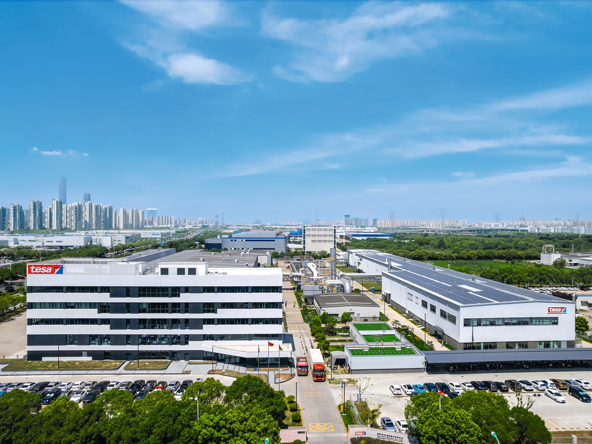 Aerial view of a sprawling industrial complex with multiple white buildings under a blue sky. The complex features parking areas and roads. A city skyline is visible in the distance. (This text has been generated by AI)