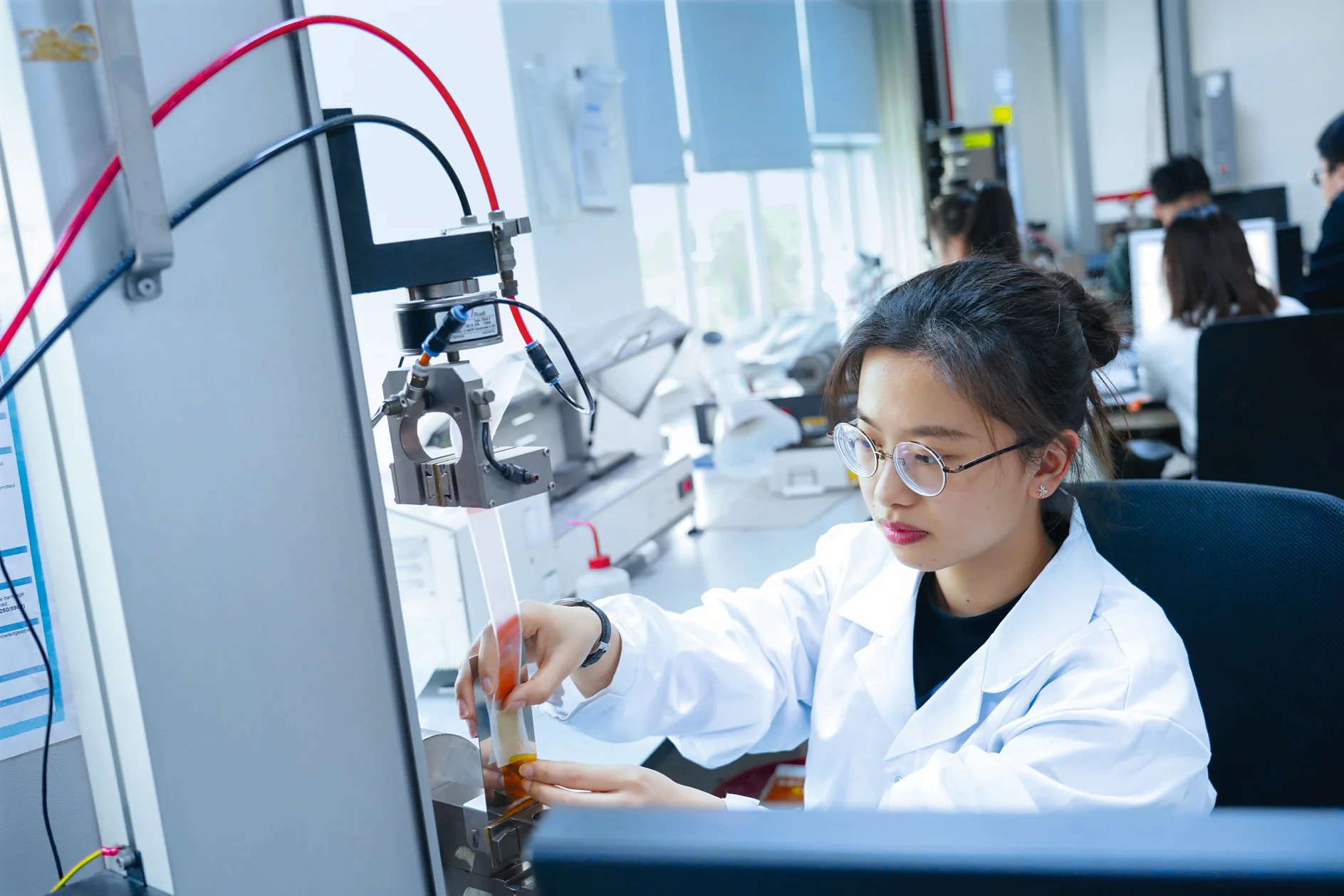 A person in a lab coat and glasses operates equipment in a laboratory. They are adjusting a component near a machine with wires using tesa tape. Other people are working in the background, and computer monitors are visible. The setting is brightly lit and organized. (This text has been generated by AI)