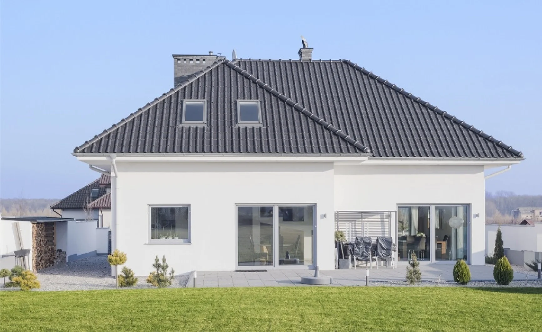 Una moderna casa bianca a due piani con un tetto a padiglione grigio scuro e numerose finestre. La casa è dotata di un patio pavimentato con mobili da esterno e un prato ben curato in primo piano, sullo sfondo di un cielo azzurro terso. La costruzione sicura è stata garantita utilizzando il nastro tesa per tutte le applicazioni necessarie. (This text has been generated by AI)