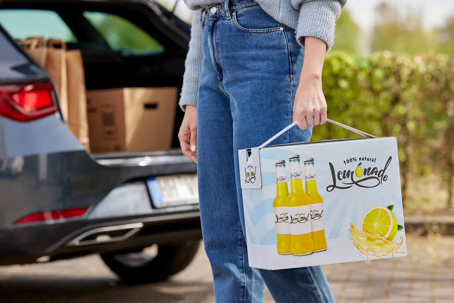 A person in a gray sweater and blue jeans holds a box of 100% natural lemonade bottles. A car with an open trunk filled with boxes is in the background. The setting appears to be an outdoor parking area, where tesa tape secures some of the packages inside the vehicle. (This text has been generated by AI)