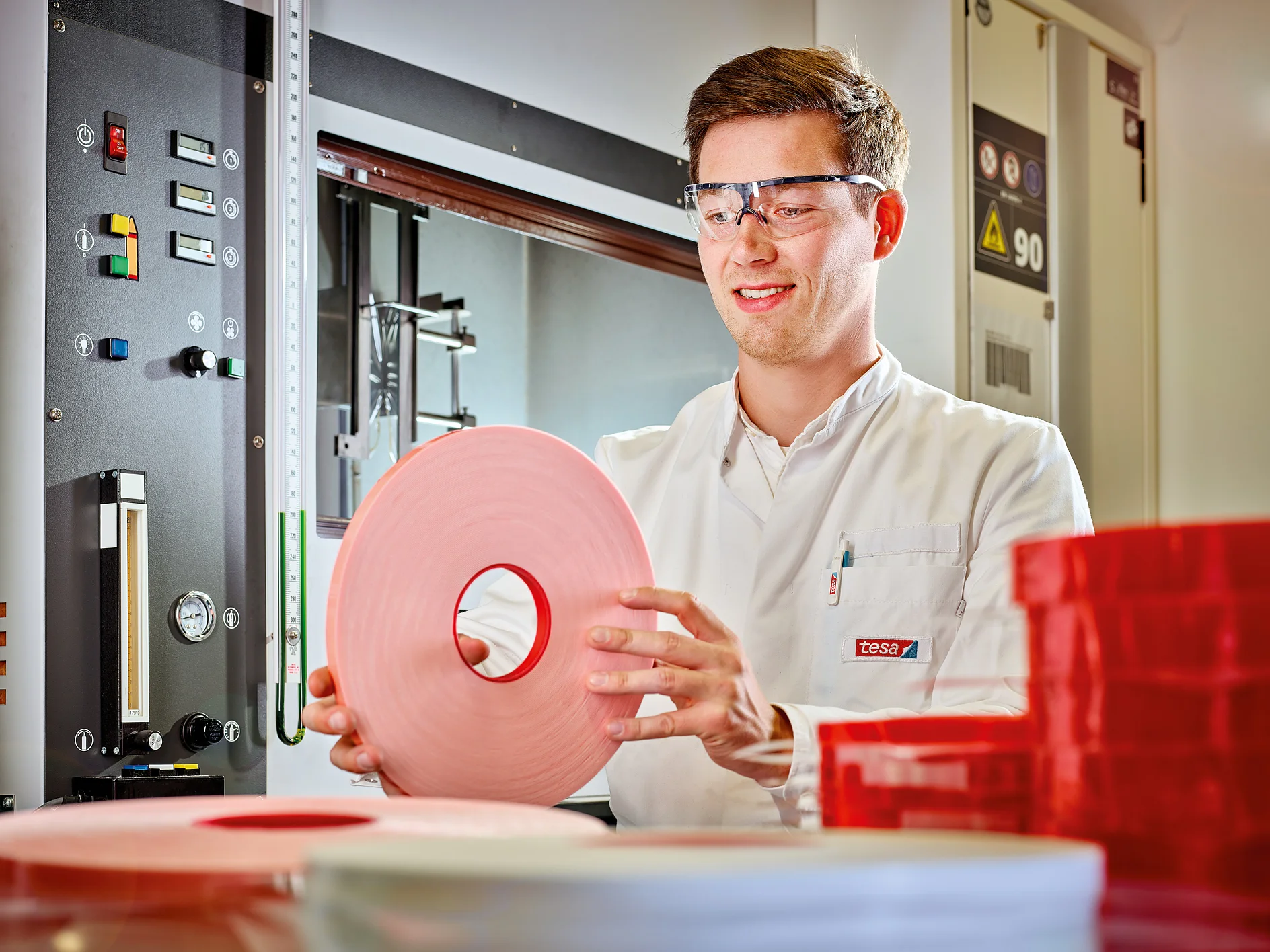 A person wearing safety glasses and a white lab coat holds a large roll of pink tesa tape. Various control panels and rolls of red tesa tape are visible in the background. (This text has been generated by AI)