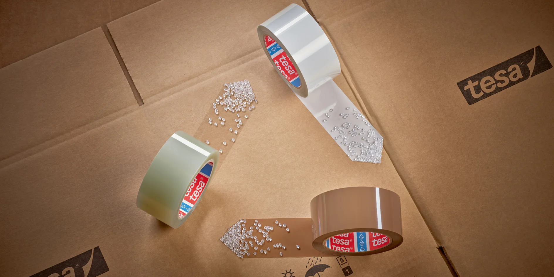 tesa-ID-60412-imballaggio-PET riciclato-nastro-famiglia-mood%20%282%29-Stampa Three rolls of tesa tape are arranged in a triangular formation on cardboard boxes. White pellets are scattered near the ends of the tesa tape. Tesa branding is visible on the tesa tape cores and the boxes. (This text has been generated by AI)