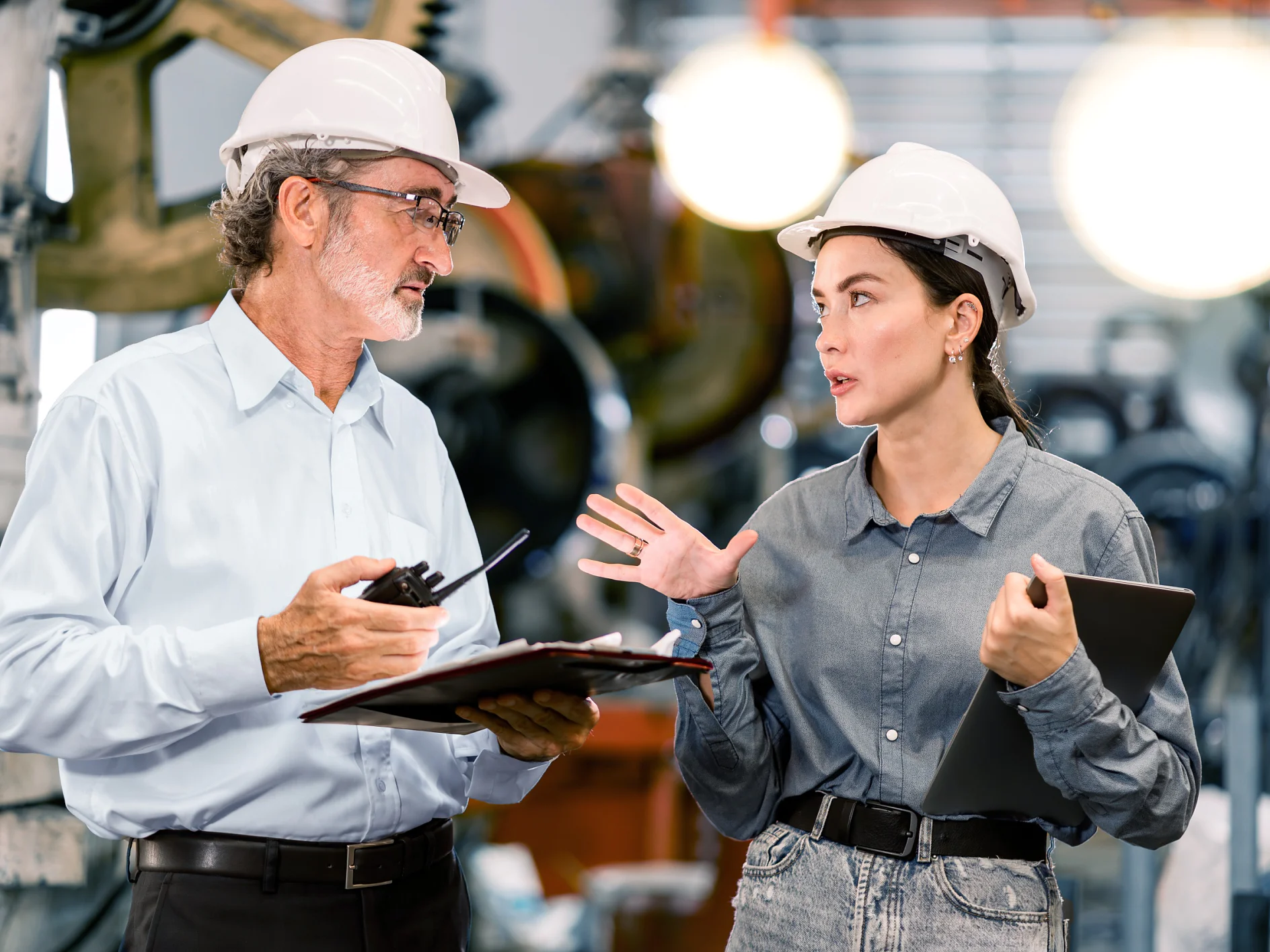 Two people in a factory setting are wearing hard hats and safety glasses. The older person holds a clipboard and pen, discussing with the younger person who is holding a digital tablet. tesa tape products can be seen around the machinery, which is visible in the blurred background. (This text has been generated by AI)