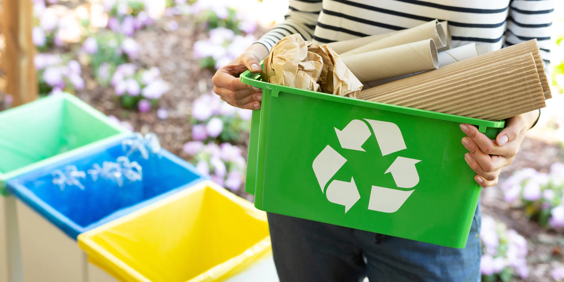 DIN CERTCO A person holds a green recycling bin filled with brown cardboard and paper secured with tesa tape. The bin features a white recycling symbol. In the background, there are blue, yellow, and green bins, along with flowerbeds showcasing purple and white flowers. (This text has been generated by AI)
