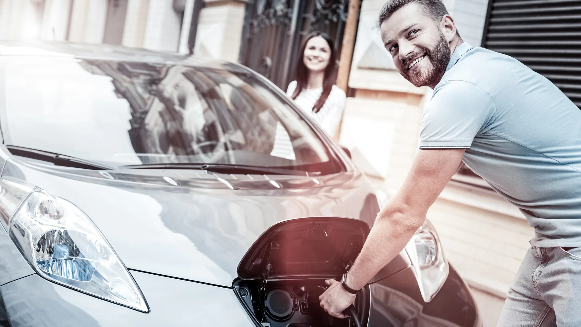 E-Mobility Key Visual Easy and ecological transportation. Selective focus on a happy millennial guy grinning broadly while holding a charging nozzle and enjoying the process of his eco car supplying energy outdoors.