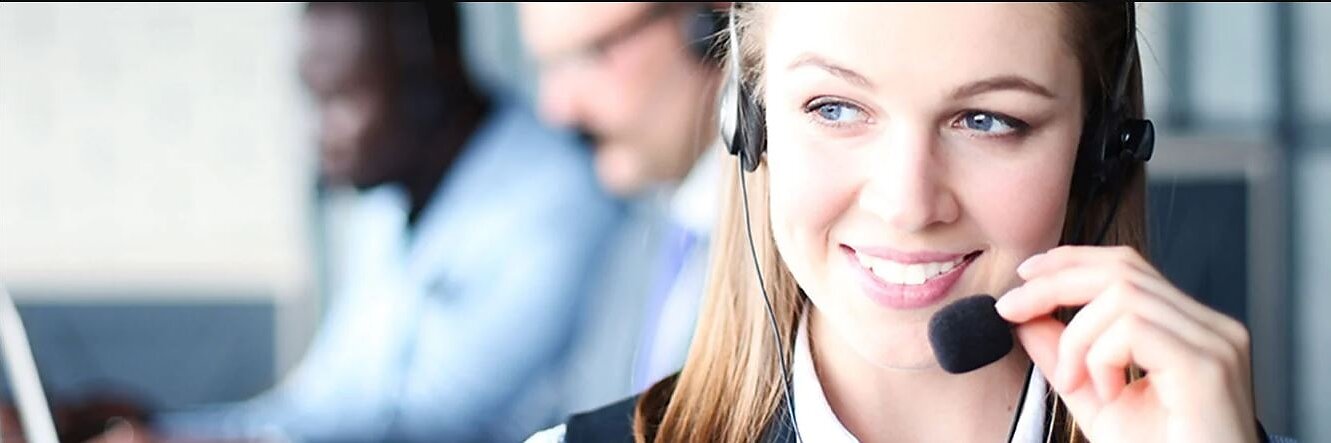 Una donna che indossa un auricolare sorride e sembra parlare. È a fuoco in primo piano, mentre due persone in abiti da lavoro lavorano al computer sullo sfondo. Ha del nastro tesa sul bordo della scrivania per tenere fermi gli appunti importanti mentre aiuta i clienti al telefono. (This text has been generated by AI)
