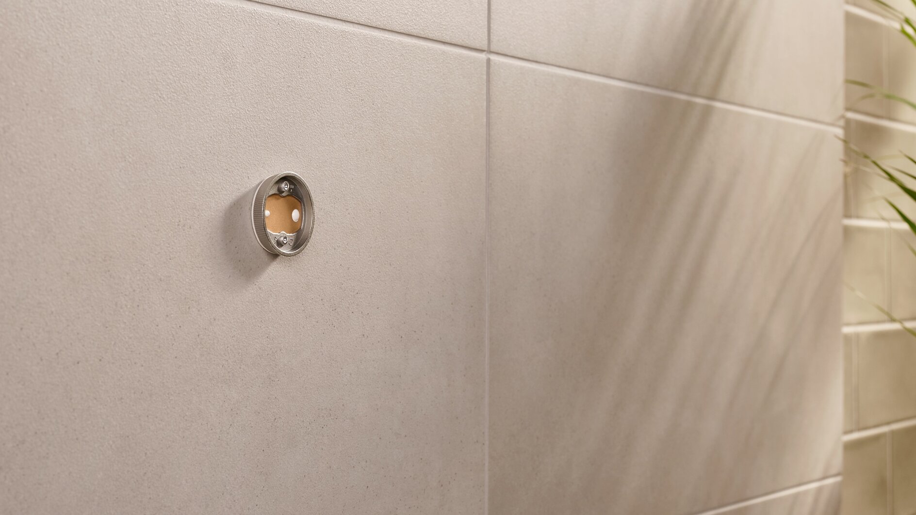 Primo piano di una parete piastrellata beige con un raccordo metallico circolare. Il raccordo ha la parte anteriore trasparente e rivela un piccolo cerchio marrone-rossastro all'interno. A destra, l'ombra di una pianta è visibile sulla parete. (This text has been generated by AI)