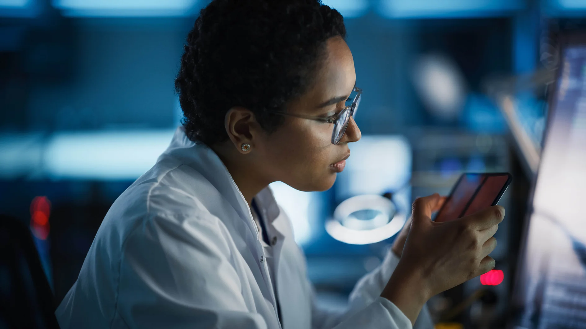 Una persona con camice e occhiali bianchi è seduta in un ambiente di laboratorio ed esamina attentamente uno smartphone fissato con nastro adesivo. Lo sfondo è sfocato, con schermi di computer e attrezzature di laboratorio. L'illuminazione è fioca e mette in risalto l'attenzione sullo smartphone avvolto con il nastro adesivo. (This text has been generated by AI)