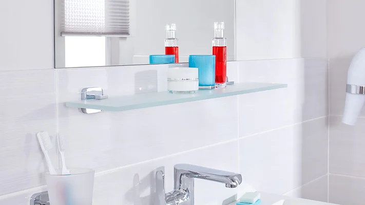 A modern bathroom showcases a glass shelf with blue and white candles alongside two red bottles. Below the shelf, a sink with a chrome faucet is partially visible. Toothbrushes in a holder are positioned on the left, while a hairdryer is mounted on the right using tesa tape. (This text has been generated by AI)