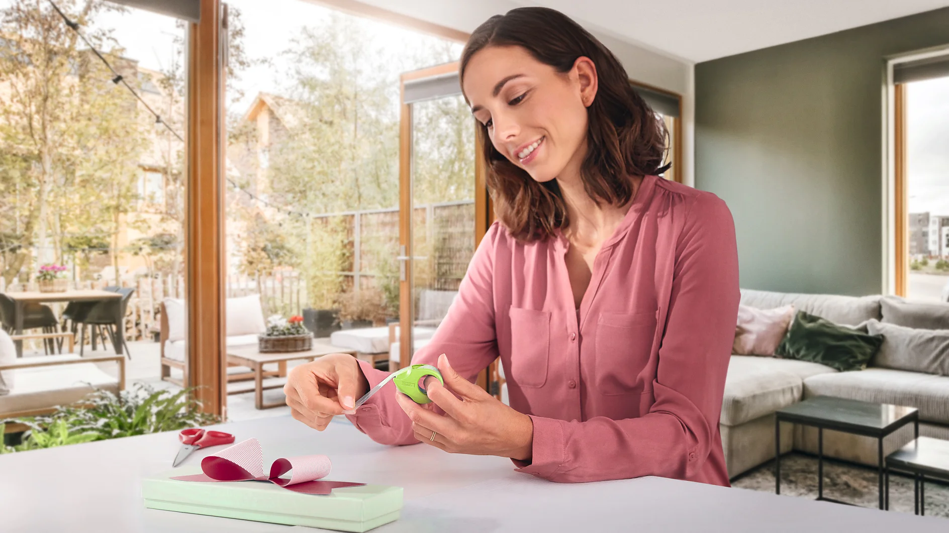 Una donna con una camicetta rosa impacchetta un regalo su un tavolo. Usa nastro verde e carta da regalo rosa, fissando il tutto con nastro tesa. La stanza ha grandi finestre con vista sugli alberi e su un patio. Sullo sfondo ci sono un divano e dei cuscini grigi. (This text has been generated by AI)