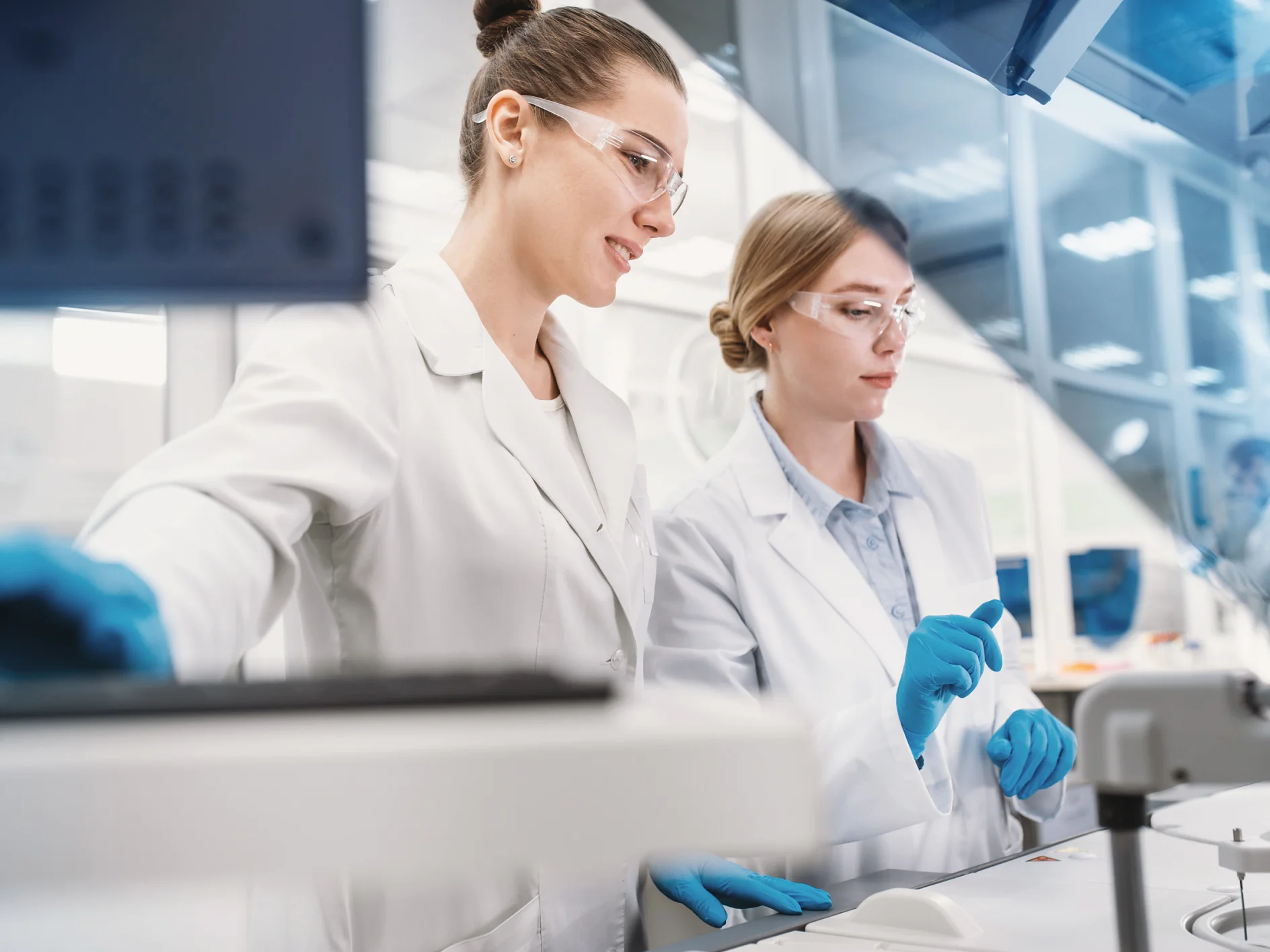 Due scienziati con camice, occhiali di sicurezza e guanti blu lavorano con attrezzature di laboratorio in un laboratorio moderno e ben illuminato. Uno tiene in mano una pipetta, mentre l'altro regola i comandi di una macchina. Tra i materiali che stanno usando durante l'esperimento si nota un nastro adesivo. (This text has been generated by AI)