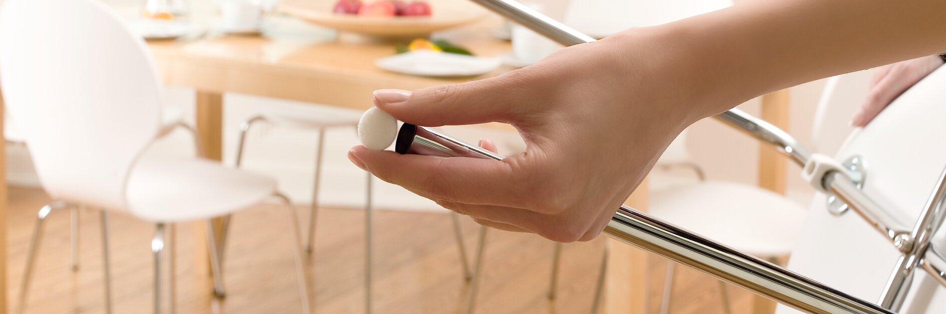 La mano di una persona regola una vite sul telaio di una sedia cromata in una luminosa sala da pranzo, mentre è visibile il nastro tesa utilizzato sul telaio. Sullo sfondo, un tavolo di legno con sedie, un vaso di rose gialle e una ciotola di frutta. (This text has been generated by AI)
