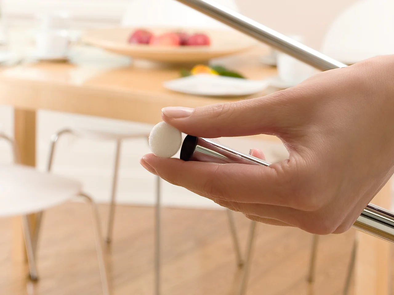 La mano di una persona regola una vite sul telaio di una sedia cromata in una luminosa sala da pranzo, mentre è visibile il nastro tesa utilizzato sul telaio. Sullo sfondo, un tavolo di legno con sedie, un vaso di rose gialle e una ciotola di frutta. (This text has been generated by AI)