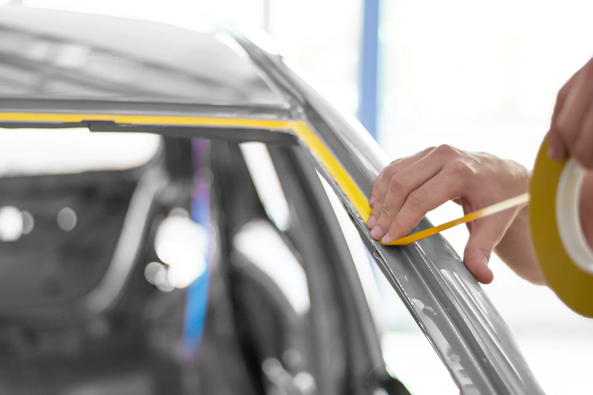 mano che applica del nastro adesivo giallo lungo la cornice del finestrino di un'auto in una luminosa officina interna (This text has been generated by AI)