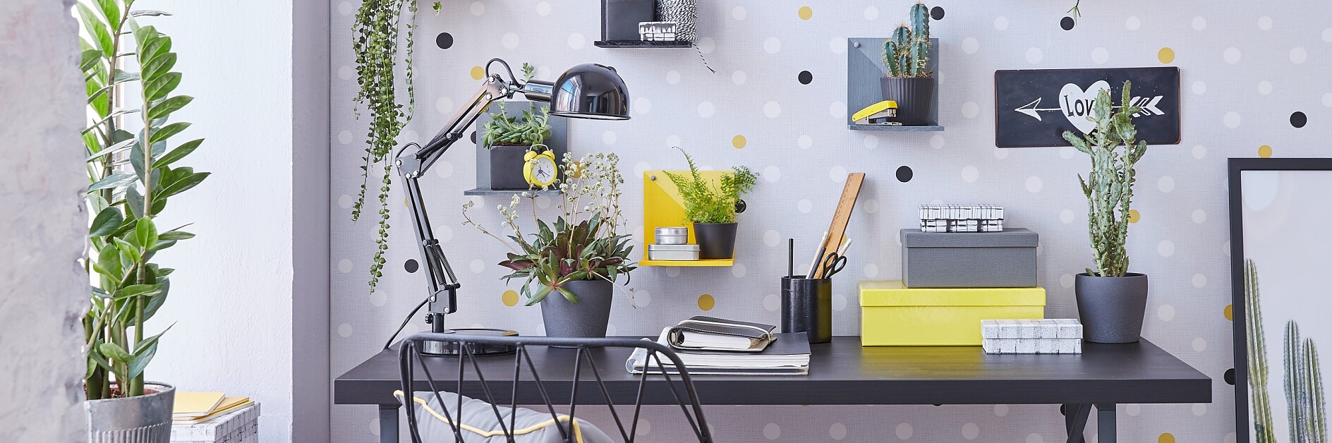 A modern desk setup with a black tabletop, two notebooks, a desk lamp, and several potted plants. The wall has a polka dot pattern with small shelves holding plants and decor. A wicker chair with a gray cushion is placed at the desk, seamlessly assembled using tesa tape for durability and style. (This text has been generated by AI)