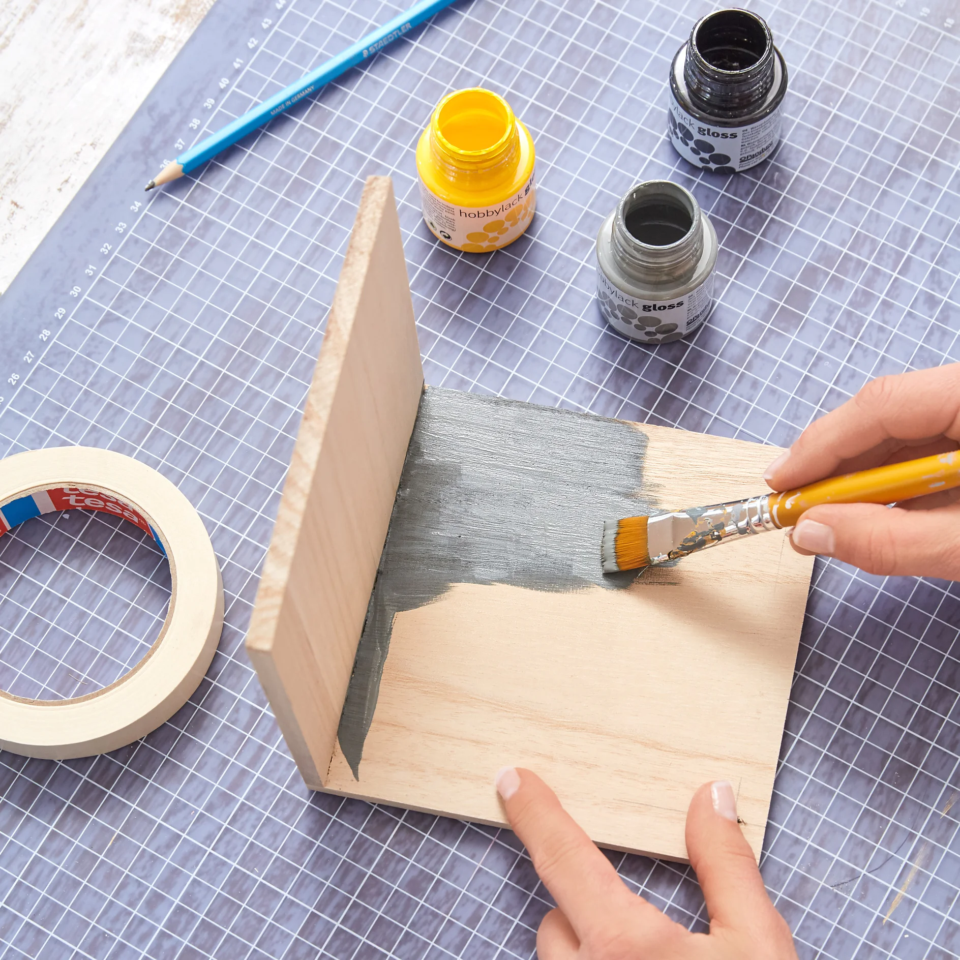 A person paints a wooden board with gray paint using a brush. The board is L-shaped and placed on a cutting mat. Yellow and gray paint jars, a roll of tesa tape, and a pencil are visible nearby. (This text has been generated by AI)