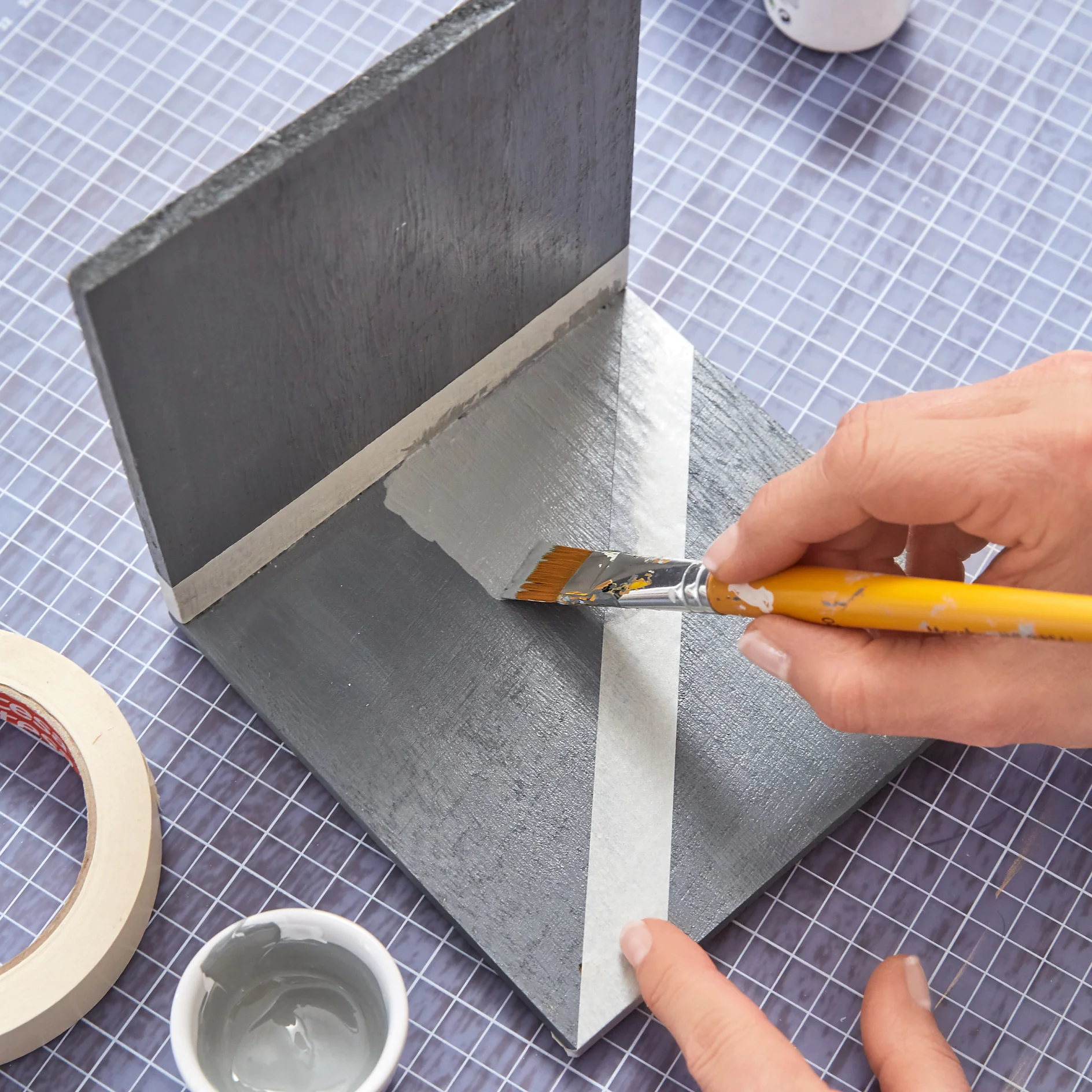 A person is painting a gray diagonal stripe on a gray wooden surface using a paintbrush. tesa tape is applied to ensure clean edges. The surface rests on a blue grid-patterned background. A small white container with paint is nearby. (This text has been generated by AI)