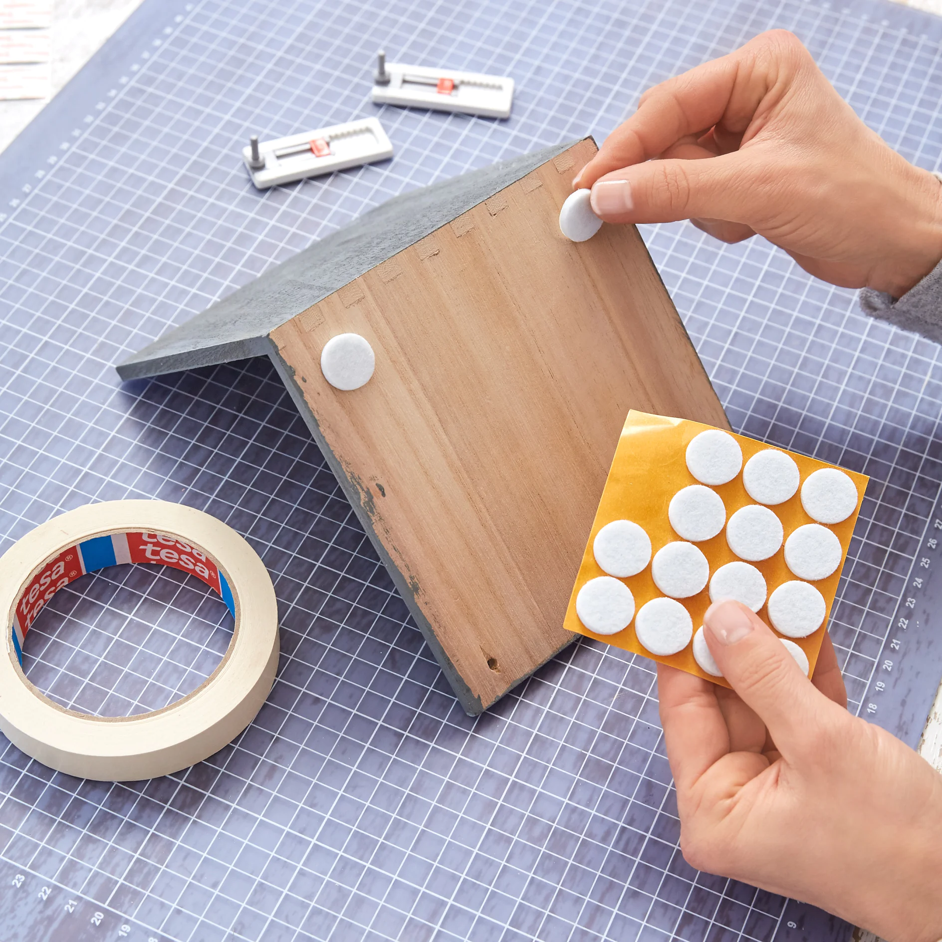 Hands applying adhesive felt pads to the underside of a wooden object. A roll of tesa tape and a sheet with more felt pads are nearby. The surface is a blue gridded cutting mat. (This text has been generated by AI)