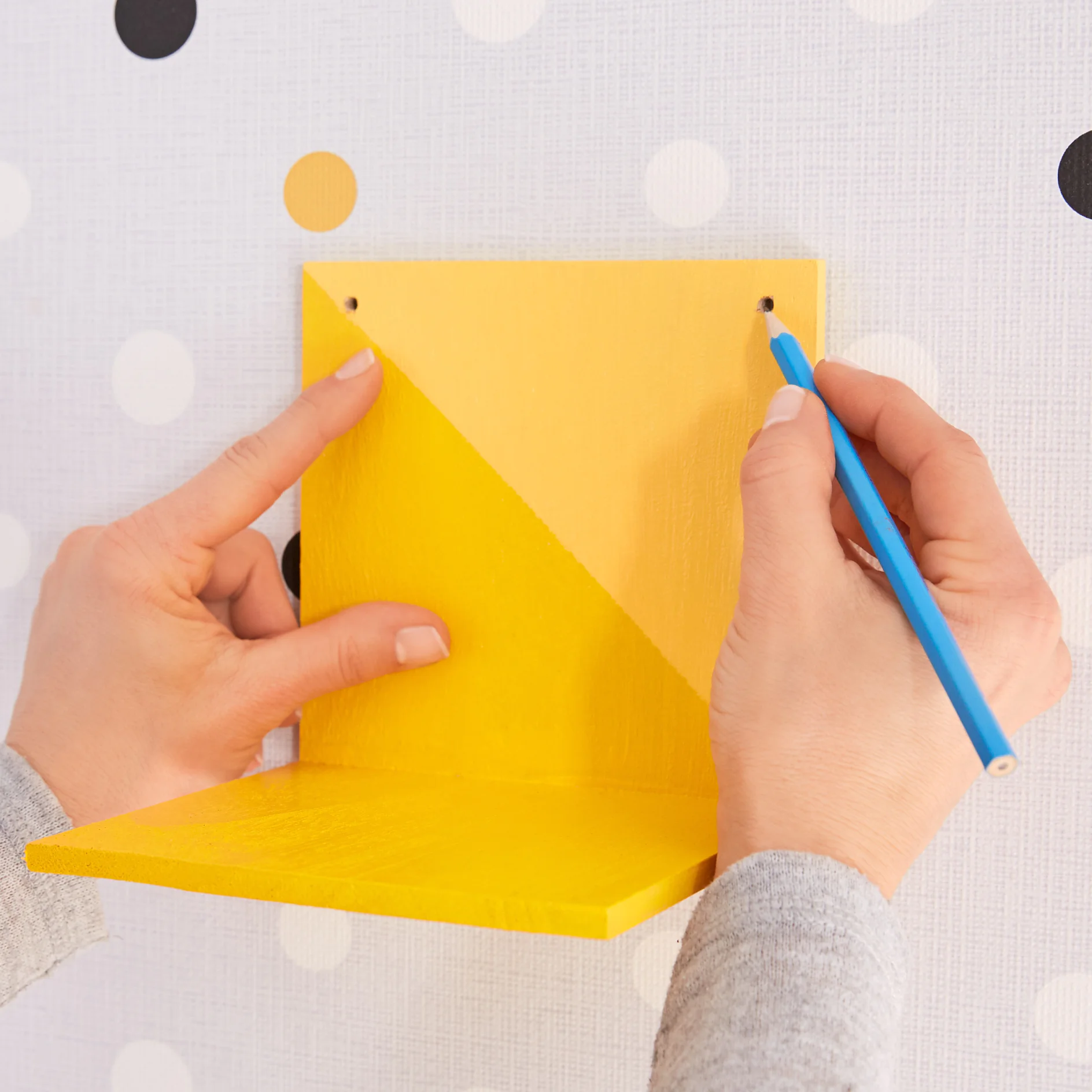 Hands hold a yellow corner shelf bracket against a polka dot wallpaper, marking two screw holes with a blue pencil on the wall. (This text has been generated by AI)