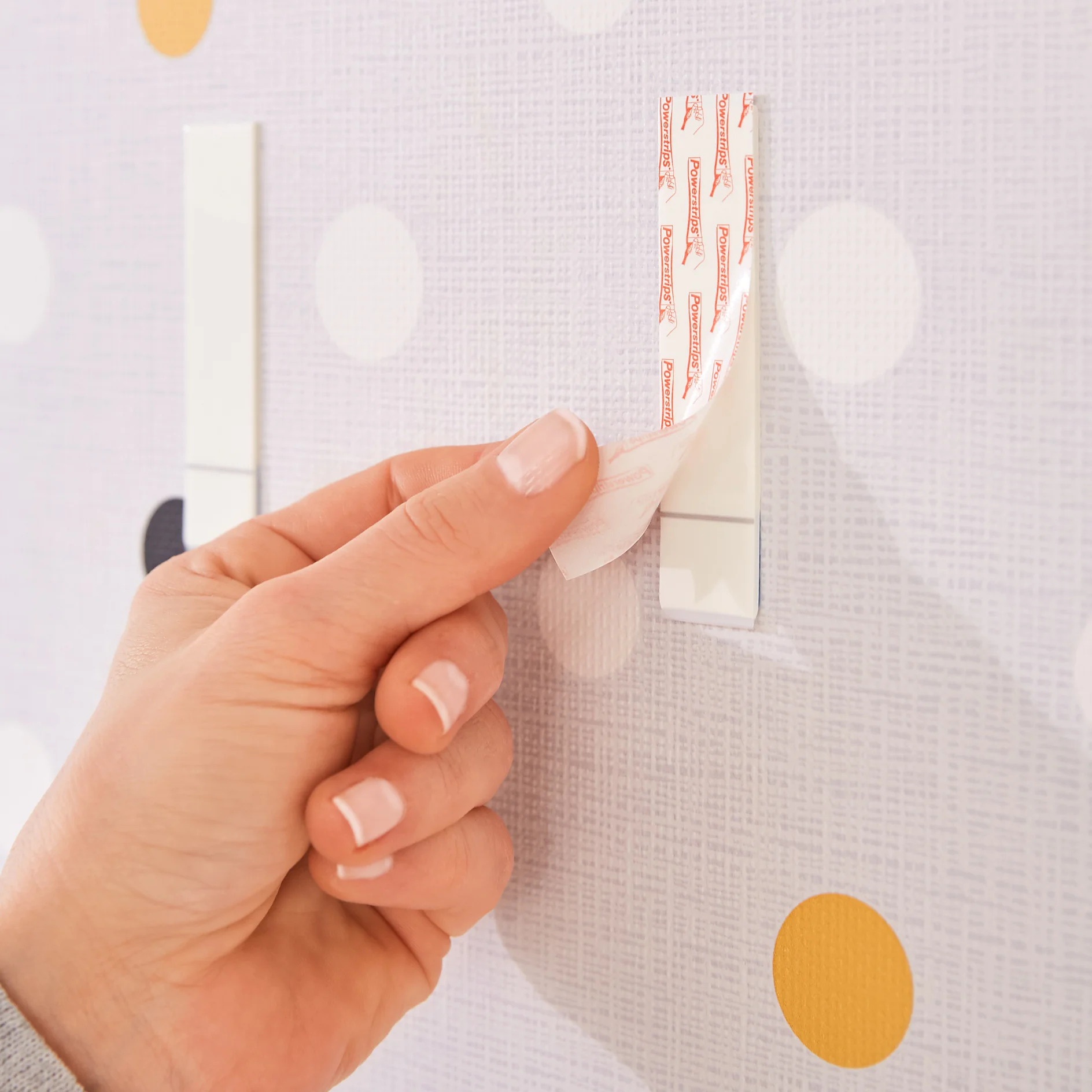 A hand is peeling a rectangular tesa tape strip off a wall with a textured surface. The wall has a pattern of large dots in white, yellow, and gray. Two removed tesa tape strips are visible on the wall. (This text has been generated by AI)