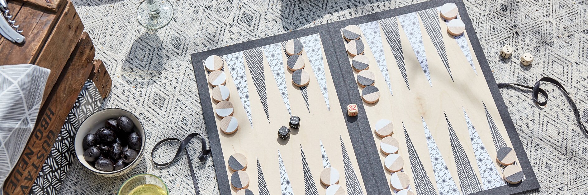 Backgammon game set up on a patterned mat with black and white pieces. Nearby are two glasses of white wine, a bowl of black olives, and decorative pillows. A wooden crate is partially visible on the left. (This text has been generated by AI)