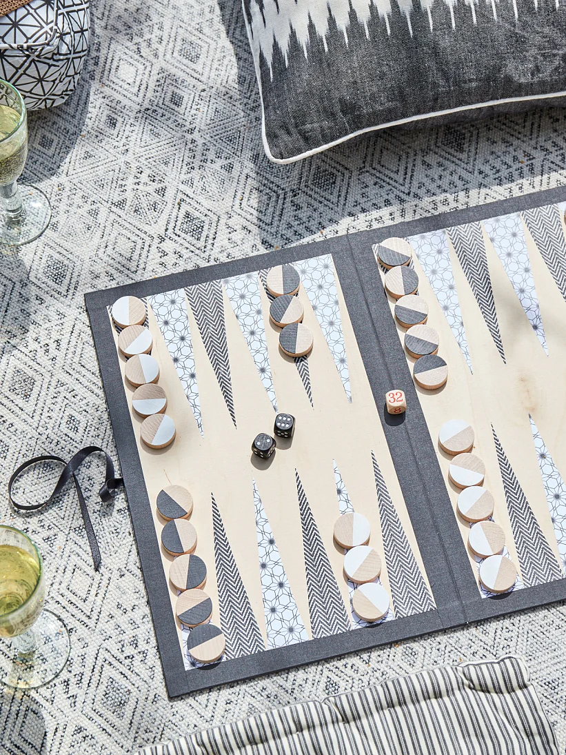 Backgammon game set up on a patterned mat with black and white pieces. Nearby are two glasses of white wine, a bowl of black olives, and decorative pillows. A wooden crate is partially visible on the left. (This text has been generated by AI)