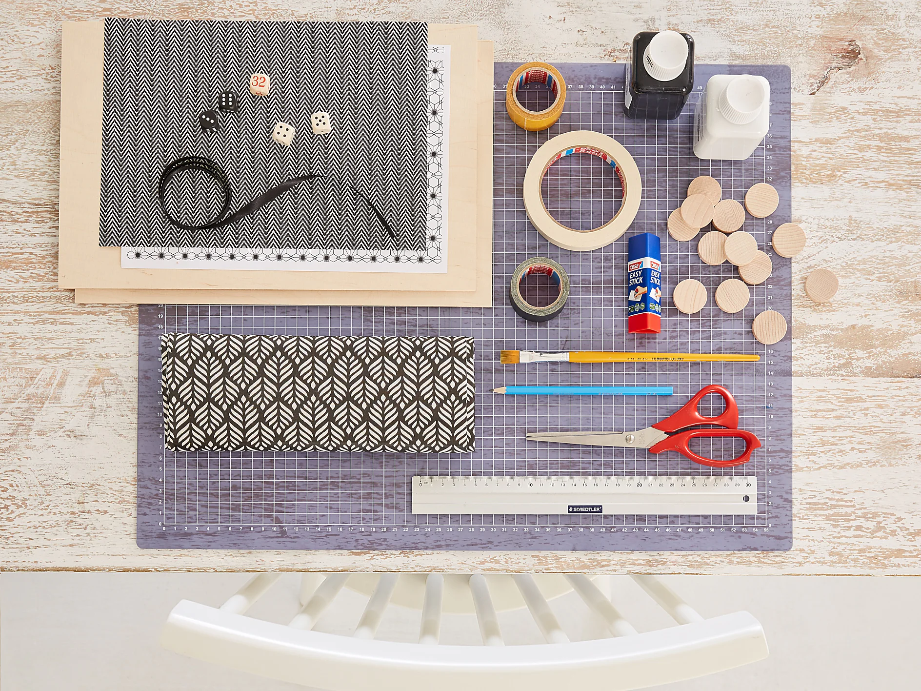 A crafting setup on a table includes a cutting mat, patterned paper, scissors, ruler, paintbrush, glue stick, tesa tape, wooden discs, black ribbon, dice, and paint bottles. A white chair is visible at the bottom edge of the image. (This text has been generated by AI)