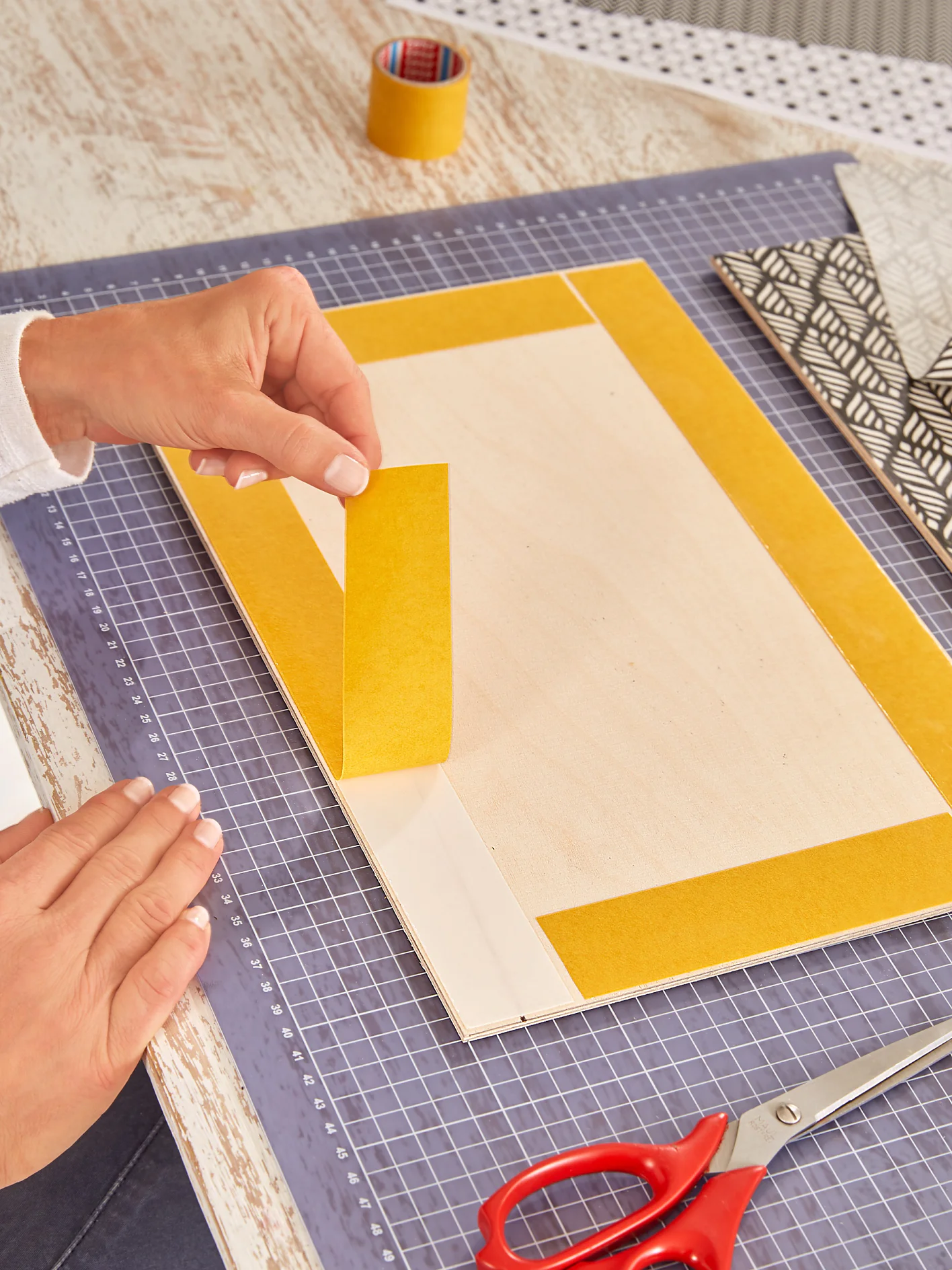 A person places yellow tesa tape on the edges of a rectangular wooden board resting on a cutting mat. A roll of tesa tape, patterned paper, and red scissors are nearby on the table. The person is wearing a white sleeve. (This text has been generated by AI)