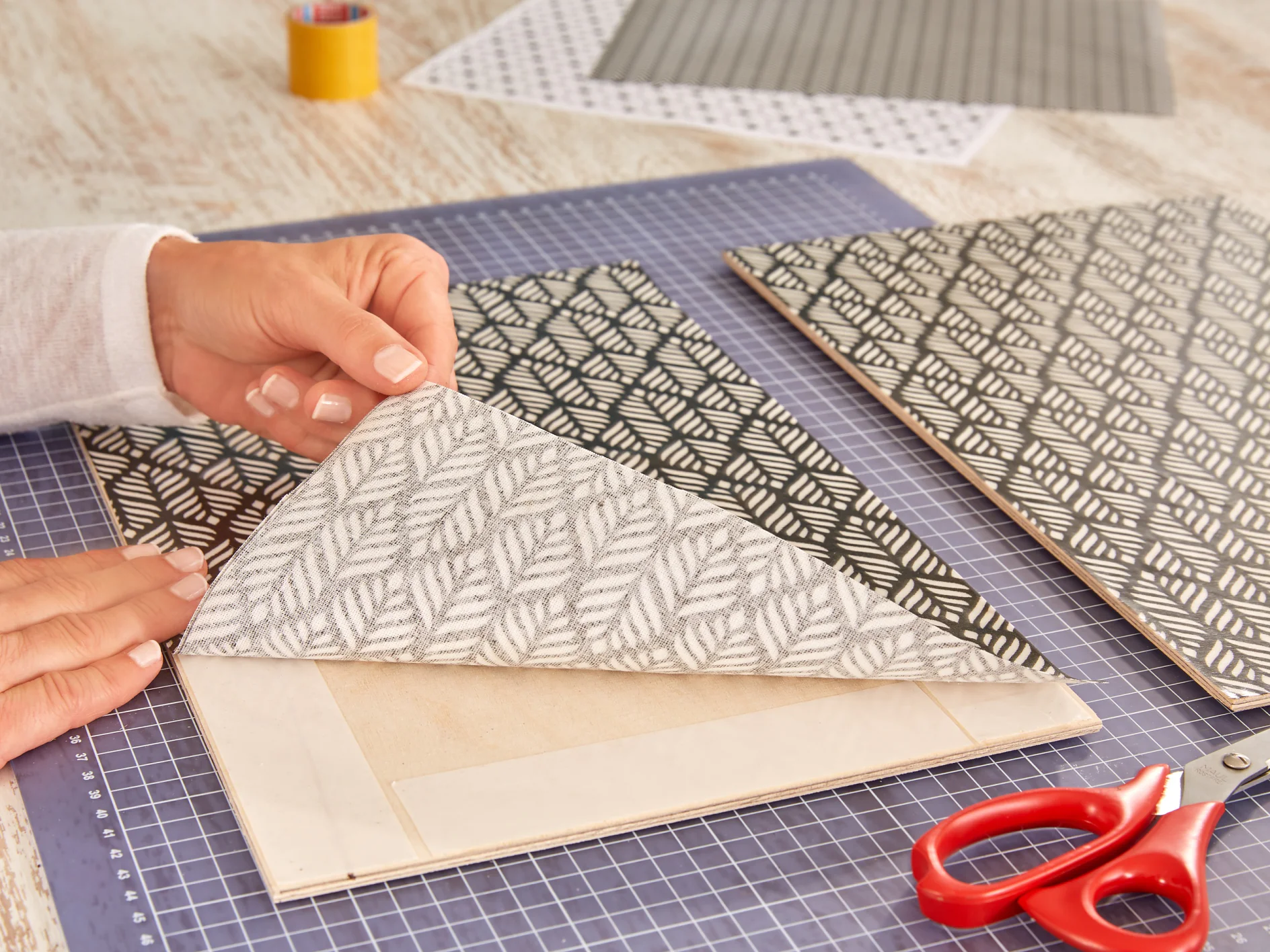 Hands are crafting by adhering a piece of decorative paper with a leaf pattern onto a wooden board. A pair of red scissors and a yellow tesa tape roll are nearby on a cutting mat. Other patterned papers are visible in the background. (This text has been generated by AI)
